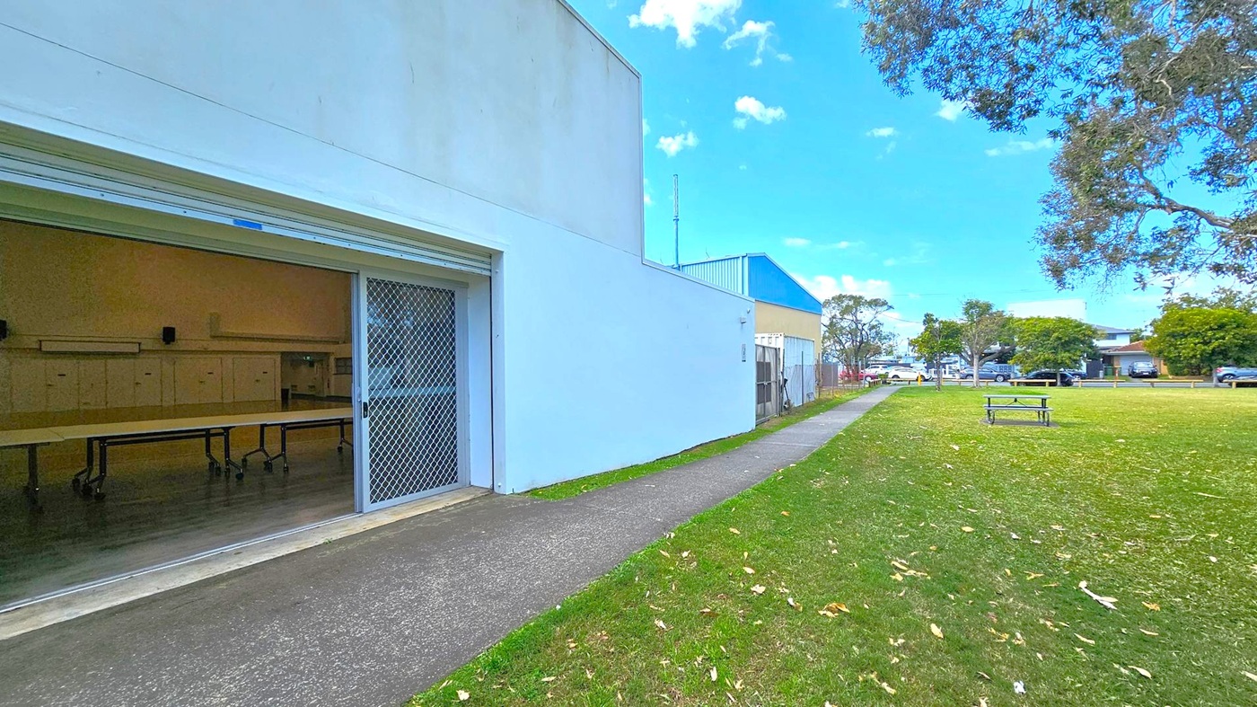 A building with a partially open roller door and a mesh screen is on the left, revealing tables inside. A concrete path runs alongside the building, leading towards the background where more buildings and parked cars are visible. On the right, a grassy area with scattered leaves and a picnic table is bordered by trees. The sky is clear and blue. A building with a partially open roller door and a mesh screen is on the left, revealing tables inside. A concrete path runs alongside the building, leading towards the background where more buildings and parked cars are visible. On the right, a grassy area with scattered leaves and a picnic table is bordered by trees. The sky is clear and blue.
