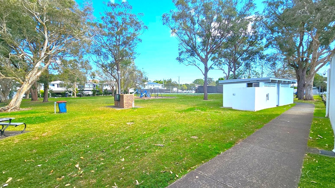 A park with a grassy area and scattered trees. On the left, there's a blue bin and a picnic table. In the centre, a playground with swings and a brick barbecue area. On the right, a white public restroom building is adjacent to a paved path. In the background, residential houses are visible beyond a fence. A park with a grassy area and scattered trees. On the left, there's a blue bin and a picnic table. In the centre, a playground with swings and a brick barbecue area. On the right, a white public restroom building is adjacent to a paved path. In the background, residential houses are visible beyond a fence.