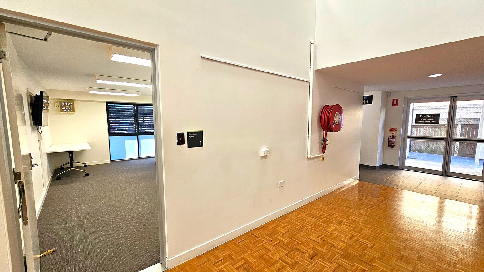 A hallway with a parquet floor and a white wall. On the left, an open door leads to a meeting room with a carpeted floor, a wall-mounted TV, and a desk. The right side features a red fire hose reel mounted on the wall. In the background, there is a fire door with a sign, and a tiled floor area leading to the outside. Ceiling lights are visible, and a sign for a meeting room is on the wall. A hallway with a parquet floor and a white wall. On the left, an open door leads to a meeting room with a carpeted floor, a wall-mounted TV, and a desk. The right side features a red fire hose reel mounted on the wall. In the background, there is a fire door with a sign, and a tiled floor area leading to the outside. Ceiling lights are visible, and a sign for a meeting room is on the wall.