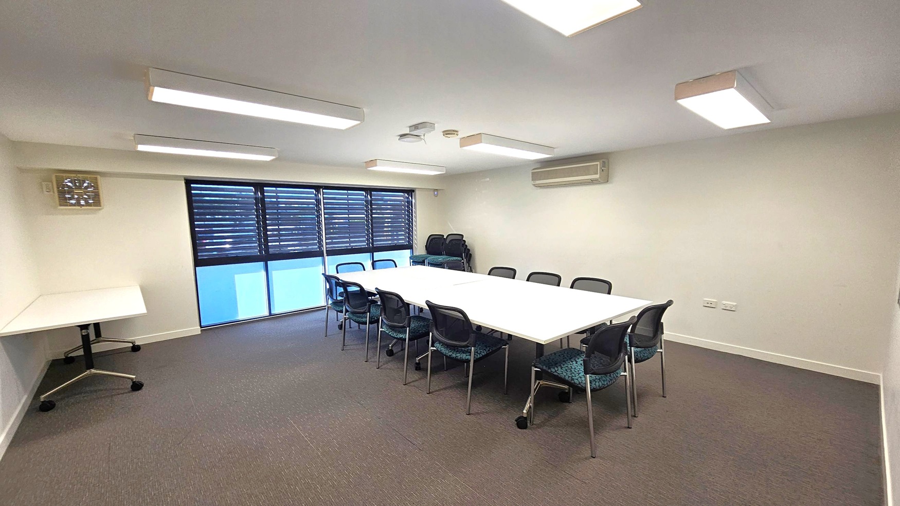 A conference room with a large rectangular table in the centre, surrounded by black chairs. On the left, there's a small table with wheels. The back wall features windows with horizontal blinds, and a stack of additional chairs is visible. The ceiling has several rectangular fluorescent lights, and an air conditioning unit is mounted on the right wall. The floor is carpeted in a grey tone. A conference room with a large rectangular table in the centre, surrounded by black chairs. On the left, there's a small table with wheels. The back wall features windows with horizontal blinds, and a stack of additional chairs is visible. The ceiling has several rectangular fluorescent lights, and an air conditioning unit is mounted on the right wall. The floor is carpeted in a grey tone.