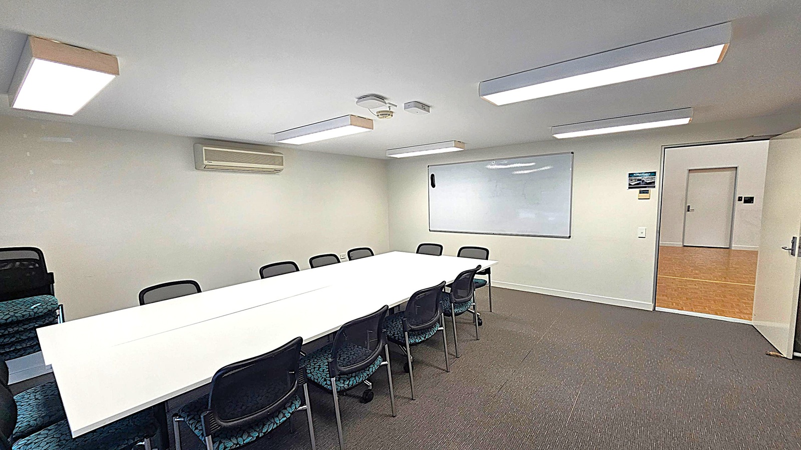 A conference room with a long white table in the centre surrounded by black chairs. On the left, there are additional stacked chairs. The back wall features a whiteboard and an air conditioning unit. The ceiling has rectangular fluorescent lights and a smoke detector. The floor is carpeted in grey. On the right, an open door leads to a hallway with a wooden floor. A conference room with a long white table in the centre surrounded by black chairs. On the left, there are additional stacked chairs. The back wall features a whiteboard and an air conditioning unit. The ceiling has rectangular fluorescent lights and a smoke detector. The floor is carpeted in grey. On the right, an open door leads to a hallway with a wooden floor.