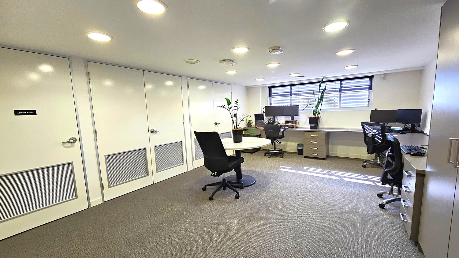 Office with grey carpet and white ceiling featuring recessed lights. On the left, two white doors, one labelled "Comms Room". In the centre, a round table with a black office chair and a potted plant. On the right, a long desk with multiple monitors, another office chair, and a potted plant. A window with horizontal blinds is above the desk. A cabinet is partially visible on the far right. Office with grey carpet and white ceiling featuring recessed lights. On the left, two white doors, one labelled "Comms Room". In the centre, a round table with a black office chair and a potted plant. On the right, a long desk with multiple monitors, another office chair, and a potted plant. A window with horizontal blinds is above the desk. A cabinet is partially visible on the far right.