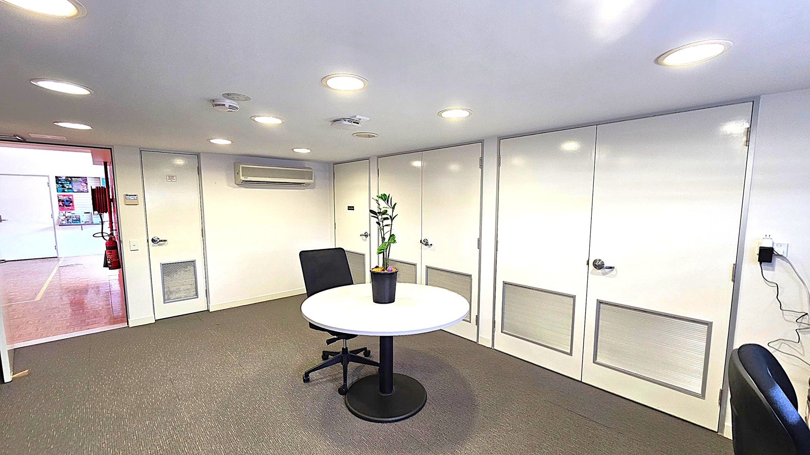 A small office space with a round table in the centre, featuring a potted plant. An office chair is positioned next to the table. The back wall has several white doors with ventilation panels and an air conditioning unit above. The floor is carpeted in a muted colour. The ceiling is fitted with recessed lights. On the left, an open doorway leads to another room with visible posters and a fire extinguisher. A power outlet with a cable is on the right wall. A small office space with a round table in the centre, featuring a potted plant. An office chair is positioned next to the table. The back wall has several white doors with ventilation panels and an air conditioning unit above. The floor is carpeted in a muted colour. The ceiling is fitted with recessed lights. On the left, an open doorway leads to another room with visible posters and a fire extinguisher. A power outlet with a cable is on the right wall.