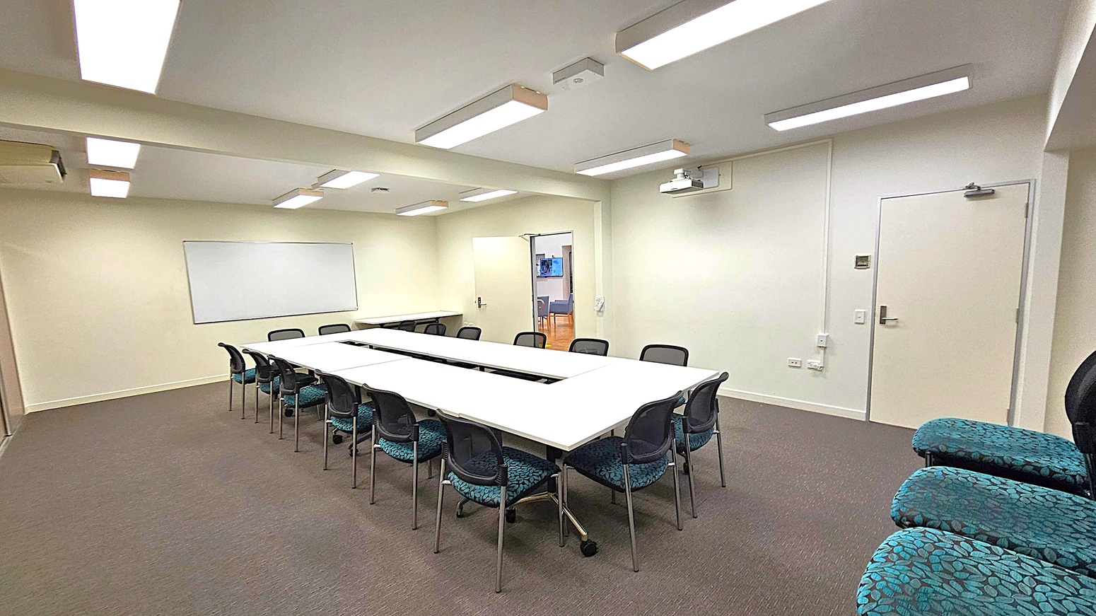 A conference room with a U-shaped arrangement of white tables surrounded by black chairs. On the left wall, a large whiteboard is mounted. The right wall has a closed door and a projector mounted on the ceiling. The floor is carpeted in grey, and the ceiling features several rectangular fluorescent lights. In the background, an open door leads to another room with visible chairs and a screen. A conference room with a U-shaped arrangement of white tables surrounded by black chairs. On the left wall, a large whiteboard is mounted. The right wall has a closed door and a projector mounted on the ceiling. The floor is carpeted in grey, and the ceiling features several rectangular fluorescent lights. In the background, an open door leads to another room with visible chairs and a screen.