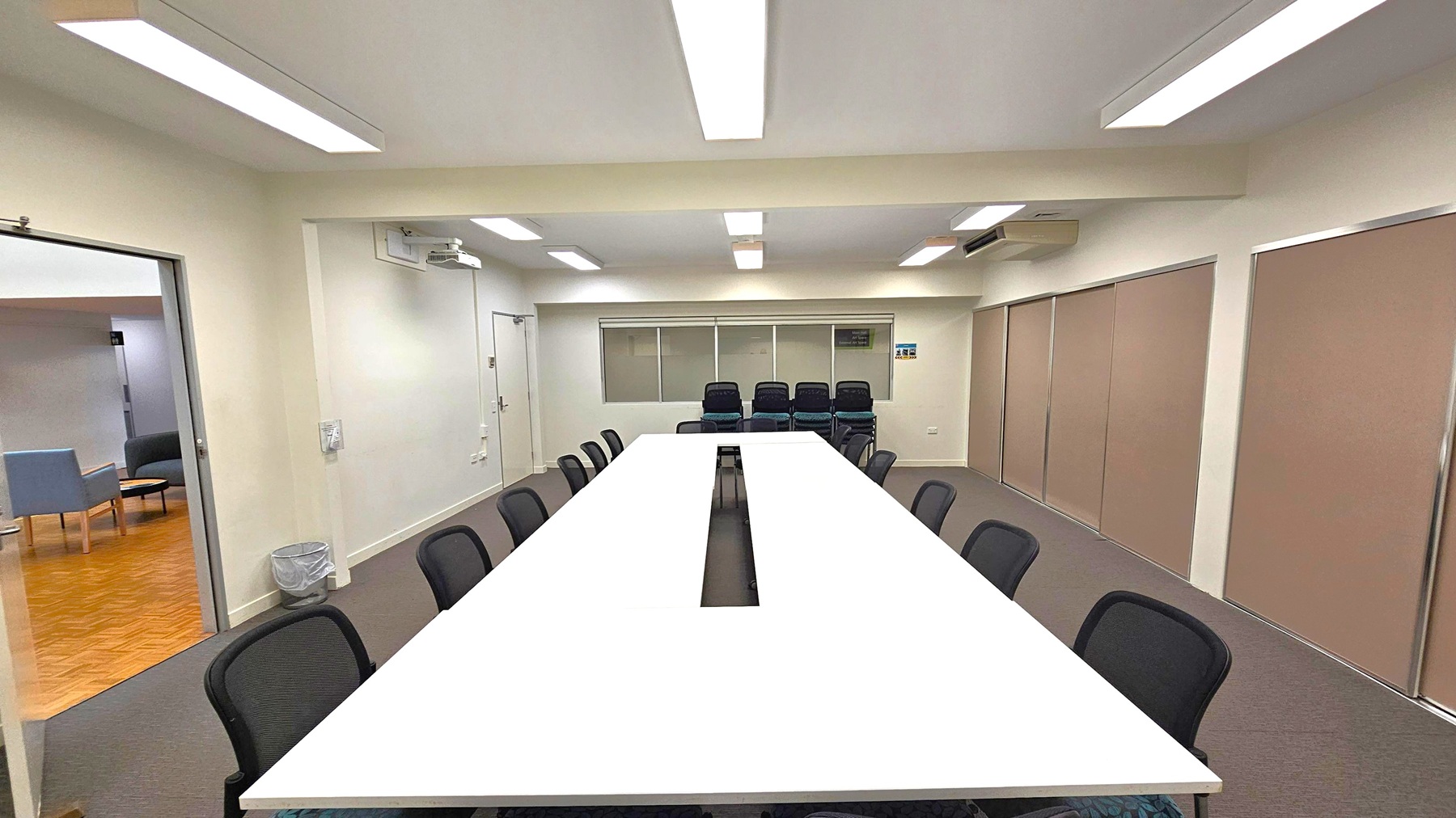 A conference room with a long, white rectangular table in the centre, surrounded by black chairs. The floor is carpeted in grey. On the left, an open door leads to another room with wooden flooring and a few chairs. The back wall has a row of windows and a stack of additional chairs. The right wall features large, beige sliding panels. The ceiling is fitted with fluorescent lights, and there is an air conditioning unit on the right wall. A conference room with a long, white rectangular table in the centre, surrounded by black chairs. The floor is carpeted in grey. On the left, an open door leads to another room with wooden flooring and a few chairs. The back wall has a row of windows and a stack of additional chairs. The right wall features large, beige sliding panels. The ceiling is fitted with fluorescent lights, and there is an air conditioning unit on the right wall.