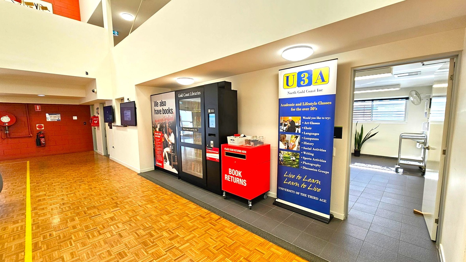 A hallway with a parquet floor and a white ceiling. On the left, a red wall with a fire hose reel and extinguisher. In the centre, a book vending machine and a red book return bin. To the right, a blue and yellow U3A banner advertising classes for those over 50. An open door leads to a room with grey tiles and a plant near the window. A hallway with a parquet floor and a white ceiling. On the left, a red wall with a fire hose reel and extinguisher. In the centre, a book vending machine and a red book return bin. To the right, a blue and yellow U3A banner advertising classes for those over 50. An open door leads to a room with grey tiles and a plant near the window.