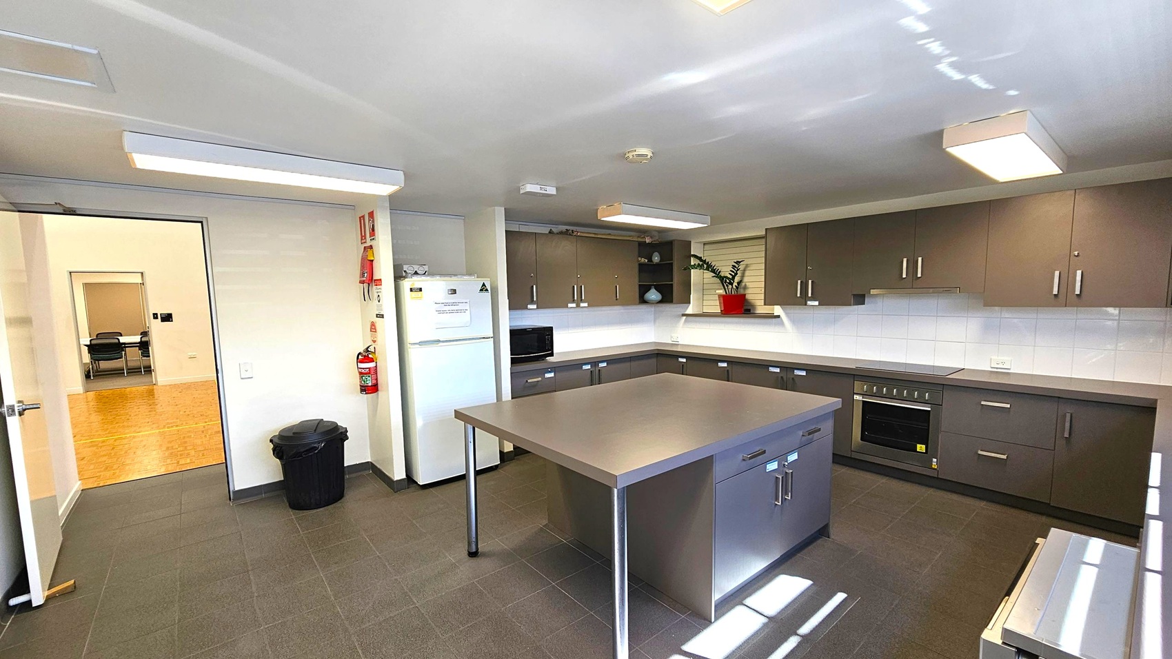 A modern kitchen with grey tiled flooring and a white ceiling featuring rectangular lights. In the centre, a large stainless steel island with drawers. The right side has dark cabinets, a built-in oven, and a white tiled backsplash. A plant in a red pot sits on the counter. On the left, a white fridge and a microwave. A fire extinguisher and a black bin are near the open door, which leads to a room with wooden flooring and chairs. A modern kitchen with grey tiled flooring and a white ceiling featuring rectangular lights. In the centre, a large stainless steel island with drawers. The right side has dark cabinets, a built-in oven, and a white tiled backsplash. A plant in a red pot sits on the counter. On the left, a white fridge and a microwave. A fire extinguisher and a black bin are near the open door, which leads to a room with wooden flooring and chairs.