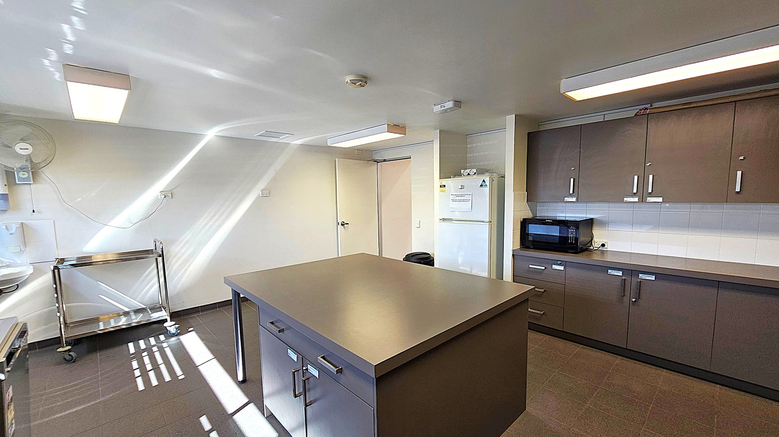 A kitchen with a central island featuring drawers and a smooth countertop. On the right, there are dark cabinets with a microwave on the counter. A white refrigerator is next to the cabinets. On the left, a metal trolley with wheels is near the wall, which has a mounted fan. The floor is tiled in a dark colour, and the ceiling has rectangular fluorescent lights. Sunlight creates patterns on the floor and walls. A kitchen with a central island featuring drawers and a smooth countertop. On the right, there are dark cabinets with a microwave on the counter. A white refrigerator is next to the cabinets. On the left, a metal trolley with wheels is near the wall, which has a mounted fan. The floor is tiled in a dark colour, and the ceiling has rectangular fluorescent lights. Sunlight creates patterns on the floor and walls.