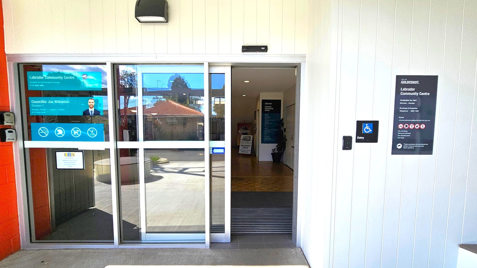 Sliding glass doors at the entrance of a community centre. On the left, signs display the centre's name and contact information, along with a photo of a person. On the right, a wall-mounted sign provides additional details about the centre, including accessibility information. A small blue sign indicates wheelchair access. Inside, a dark hallway is visible with a plant and a sign. The exterior features a light-coloured wall and a concrete floor. Sliding glass doors at the entrance of a community centre. On the left, signs display the centre's name and contact information, along with a photo of a person. On the right, a wall-mounted sign provides additional details about the centre, including accessibility information. A small blue sign indicates wheelchair access. Inside, a dark hallway is visible with a plant and a sign. The exterior features a light-coloured wall and a concrete floor.