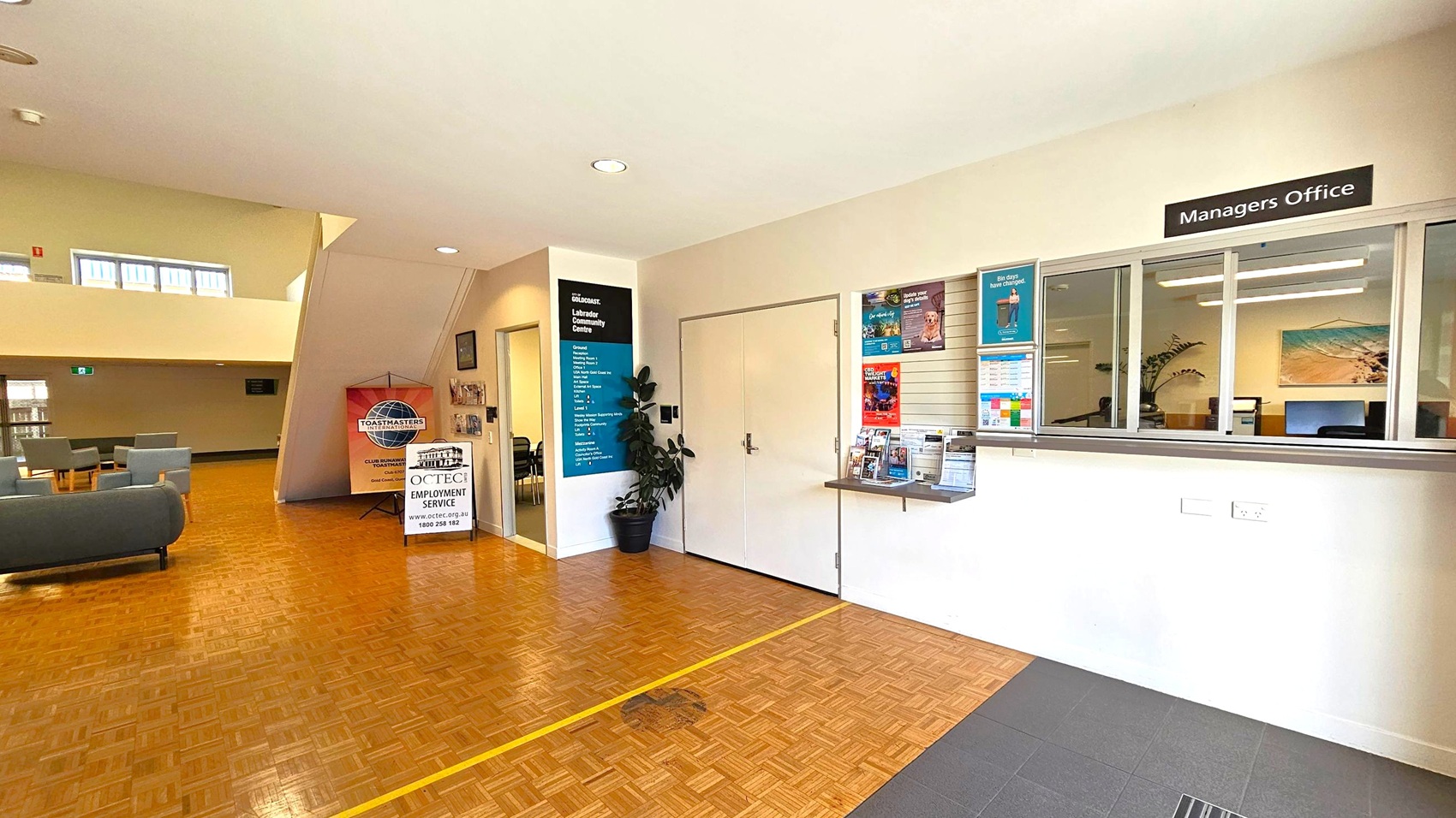 A spacious interior with a parquet floor. On the right, there's a "Managers Office" with a window and various posters. In the centre, a door is flanked by a plant and a sign listing services. On the left, a seating area with grey chairs and a table. A sign for "Toastmasters" and "CCTEC Employment Service" is visible. The ceiling is light-coloured with recessed lighting, and a staircase leads to an upper level with windows. A spacious interior with a parquet floor. On the right, there's a "Managers Office" with a window and various posters. In the centre, a door is flanked by a plant and a sign listing services. On the left, a seating area with grey chairs and a table. A sign for "Toastmasters" and "CCTEC Employment Service" is visible. The ceiling is light-coloured with recessed lighting, and a staircase leads to an upper level with windows.