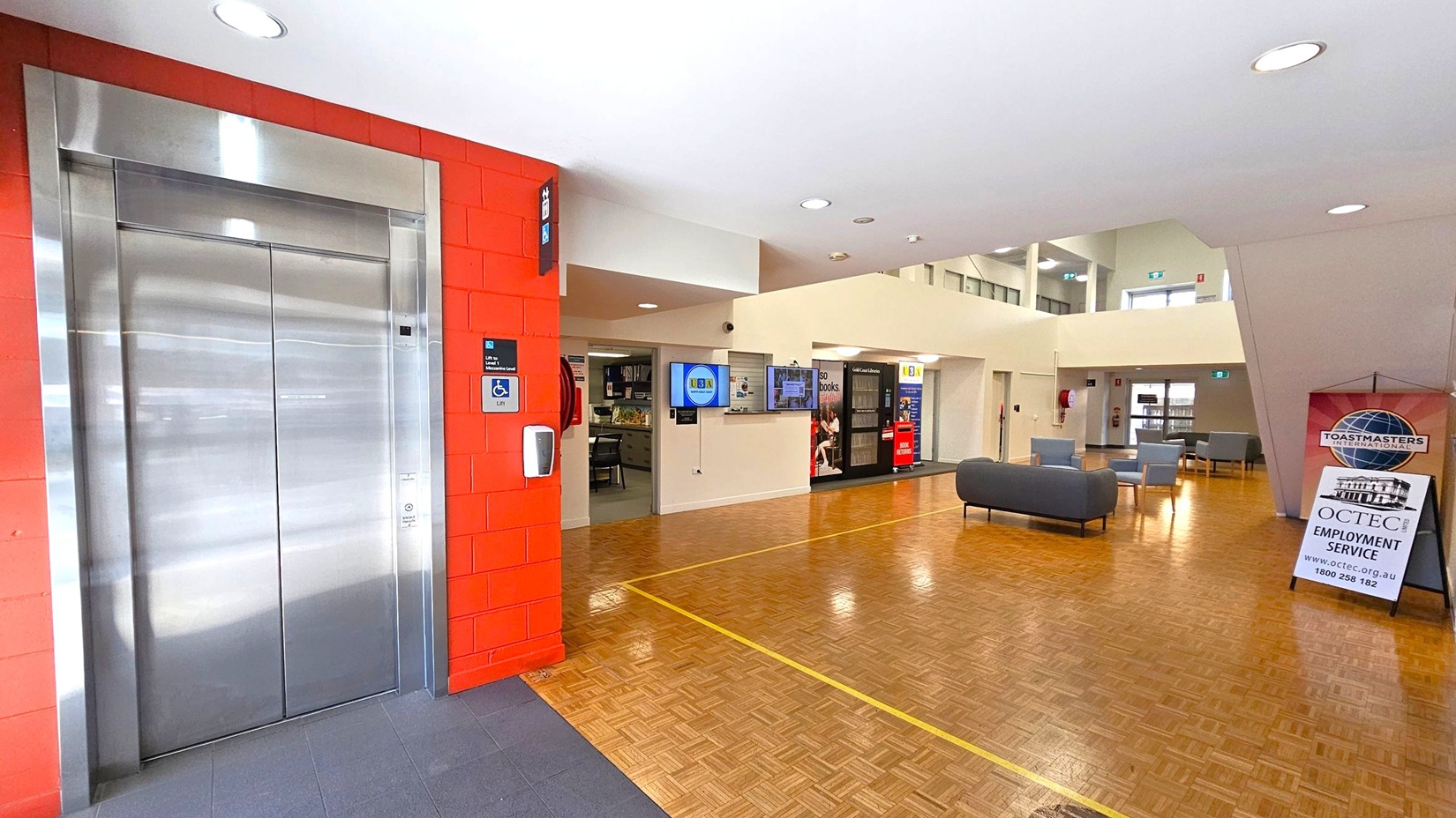 On the left, there is a stainless steel lift with a red brick wall. A sign indicates accessibility features. In the centre, a digital screen is mounted on a cream wall, with a small office area visible. The floor is wooden with a yellow line. On the right, there are grey sofas and chairs, and a sign for OCTEC Employment Service. The ceiling is white with recessed lighting. On the left, there is a stainless steel lift with a red brick wall. A sign indicates accessibility features. In the centre, a digital screen is mounted on a cream wall, with a small office area visible. The floor is wooden with a yellow line. On the right, there are grey sofas and chairs, and a sign for OCTEC Employment Service. The ceiling is white with recessed lighting.