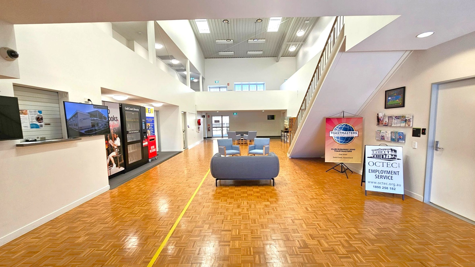 A spacious interior with a parquet floor and high ceiling. On the left, there's a counter with a TV screen and a vending machine. In the centre, a seating area with a sofa and chairs. On the right, a staircase leads to an upper level. Signs for Toastmasters International and OCTEC Employment Service are displayed near the wall, along with a brochure rack. The ceiling features skylights and recessed lighting. A spacious interior with a parquet floor and high ceiling. On the left, there's a counter with a TV screen and a vending machine. In the centre, a seating area with a sofa and chairs. On the right, a staircase leads to an upper level. Signs for Toastmasters International and OCTEC Employment Service are displayed near the wall, along with a brochure rack. The ceiling features skylights and recessed lighting.