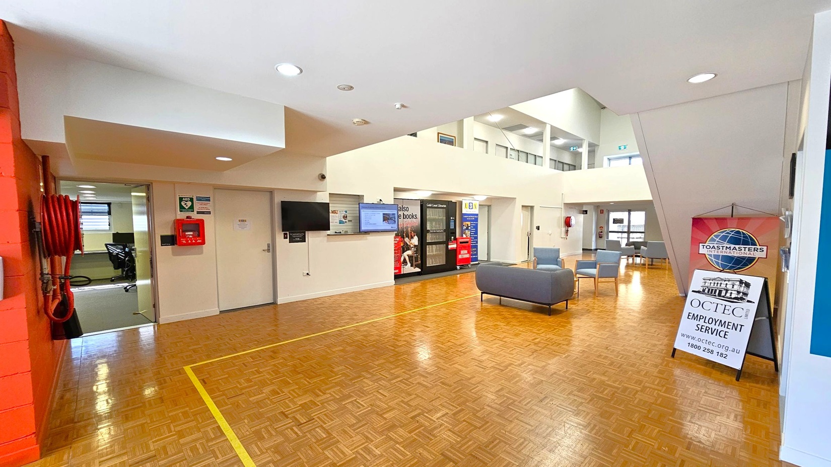A spacious interior with a parquet wooden floor and a white ceiling with recessed lights. On the left, a red fire hose and a partially open door leading to an office area. In the centre, a wall-mounted TV and a vending machine. On the right, a seating area with grey chairs and a sign for Toastmasters International and OCTEC Employment Service. The background features large windows and additional seating. A spacious interior with a parquet wooden floor and a white ceiling with recessed lights. On the left, a red fire hose and a partially open door leading to an office area. In the centre, a wall-mounted TV and a vending machine. On the right, a seating area with grey chairs and a sign for Toastmasters International and OCTEC Employment Service. The background features large windows and additional seating.