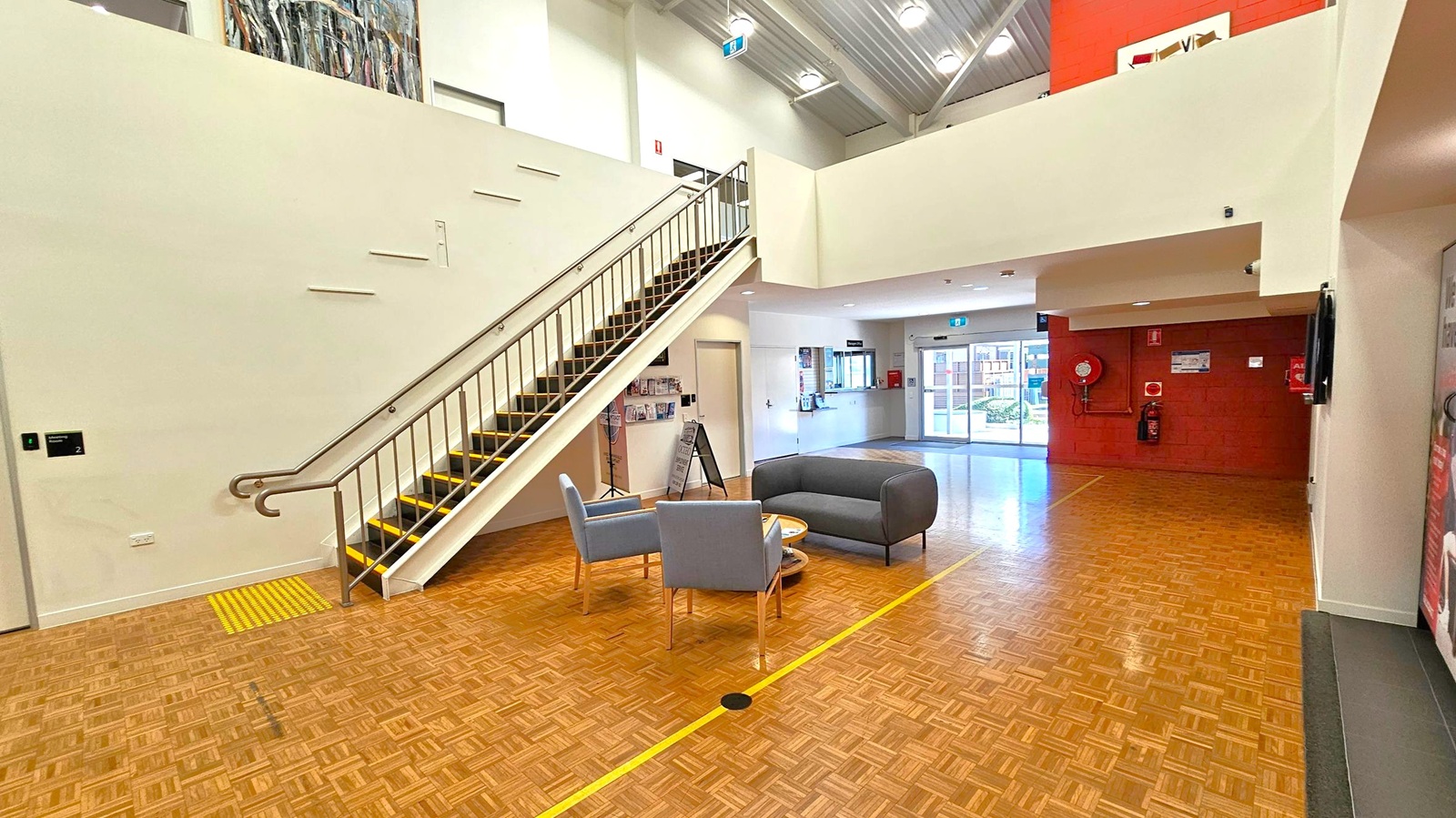 Spacious interior with a parquet floor. On the left, a staircase with a metal railing leads to an upper level. Below the stairs, a wall-mounted display holds brochures. In the centre, a seating area with two grey chairs and a small table. The right side features a red wall with a fire extinguisher and a hose reel. The background shows a reception area with a window and a glass door leading outside. The ceiling is high with exposed beams and lights. Spacious interior with a parquet floor. On the left, a staircase with a metal railing leads to an upper level. Below the stairs, a wall-mounted display holds brochures. In the centre, a seating area with two grey chairs and a small table. The right side features a red wall with a fire extinguisher and a hose reel. The background shows a reception area with a window and a glass door leading outside. The ceiling is high with exposed beams and lights.