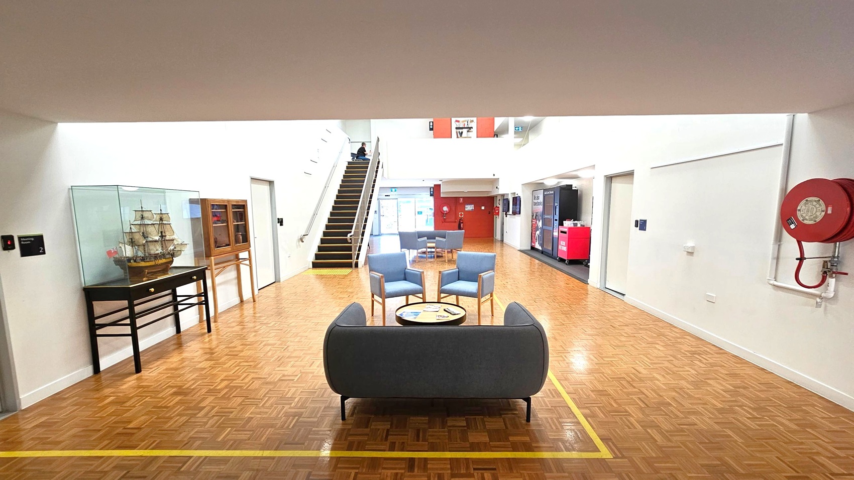 A spacious indoor area with a parquet floor. In the foreground, a seating arrangement includes a grey sofa and three blue chairs around a circular table. On the left, a model ship is displayed in a glass case on a black table. A staircase leads up to the left, and a fire hose reel is mounted on the right wall. In the background, vending machines and a red wall are visible, with more seating near a glass entrance. The ceiling is plain and white. A spacious indoor area with a parquet floor. In the foreground, a seating arrangement includes a grey sofa and three blue chairs around a circular table. On the left, a model ship is displayed in a glass case on a black table. A staircase leads up to the left, and a fire hose reel is mounted on the right wall. In the background, vending machines and a red wall are visible, with more seating near a glass entrance. The ceiling is plain and white.