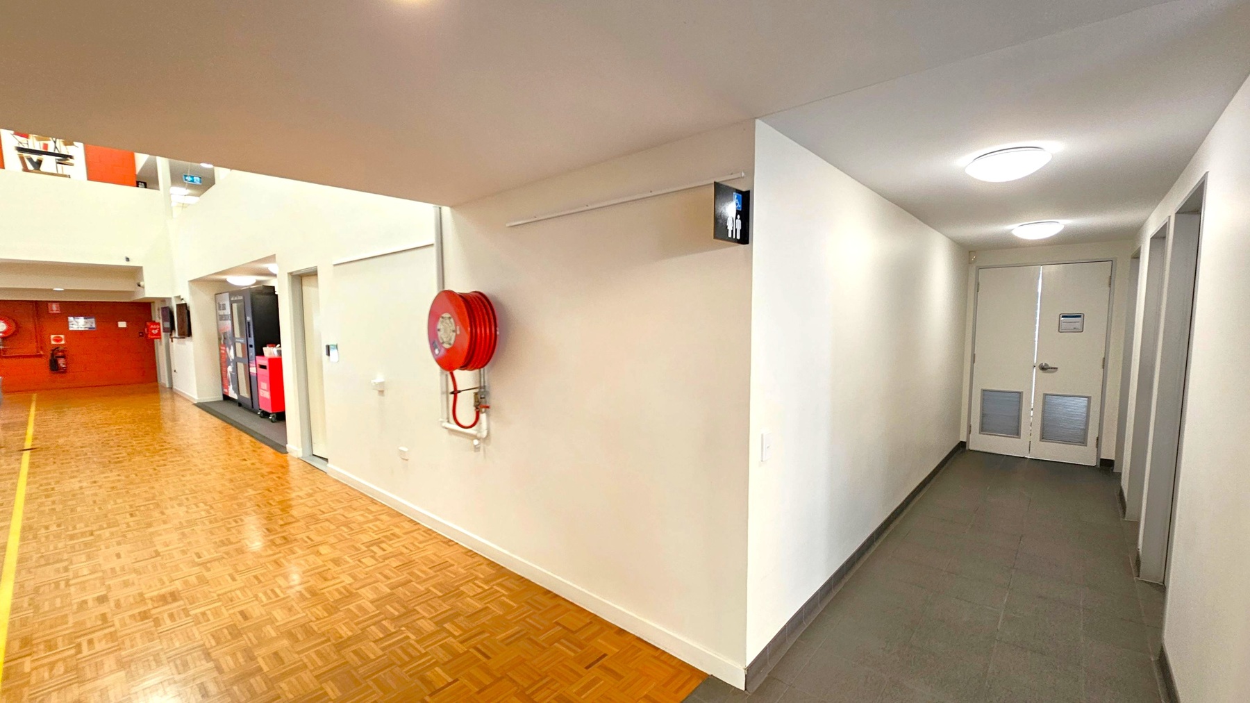 A hallway with a wooden parquet floor on the left and a tiled floor on the right. A red fire hose reel is mounted on the wall near the corner. A sign with male and female figures indicates a restroom down the corridor on the right. The ceiling is white with circular lights. In the background, there are vending machines and a red wall with fire safety equipment. A hallway with a wooden parquet floor on the left and a tiled floor on the right. A red fire hose reel is mounted on the wall near the corner. A sign with male and female figures indicates a restroom down the corridor on the right. The ceiling is white with circular lights. In the background, there are vending machines and a red wall with fire safety equipment.