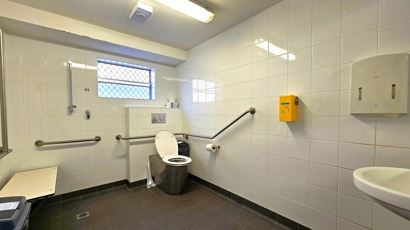 A public restroom with white tiled walls and a dark tiled floor. In the centre, a toilet with a raised seat and a lid is flanked by metal grab bars. Above the toilet, a small window with a wire mesh cover allows light in. To the right, a yellow sharps disposal box and a soap dispenser are mounted on the wall. A sink is partially visible in the foreground on the right. On the left, a foldable bench and a waste bin are present. The ceiling has a fluorescent light fixture. A public restroom with white tiled walls and a dark tiled floor. In the centre, a toilet with a raised seat and a lid is flanked by metal grab bars. Above the toilet, a small window with a wire mesh cover allows light in. To the right, a yellow sharps disposal box and a soap dispenser are mounted on the wall. A sink is partially visible in the foreground on the right. On the left, a foldable bench and a waste bin are present. The ceiling has a fluorescent light fixture.