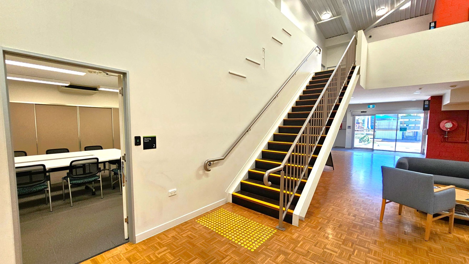 On the left, there's an open door leading to a meeting room with a long table and several chairs. The floor is carpeted inside the room. In the centre, a staircase with black steps and yellow edges ascends to the upper level, with a metal handrail on the right. The floor is wooden parquet. On the right, there's a seating area with grey chairs and a table. In the background, large glass doors provide a view of the outside, and a red wall features a fire hose reel. The ceiling is high with recessed lighting. On the left, there's an open door leading to a meeting room with a long table and several chairs. The floor is carpeted inside the room. In the centre, a staircase with black steps and yellow edges ascends to the upper level, with a metal handrail on the right. The floor is wooden parquet. On the right, there's a seating area with grey chairs and a table. In the background, large glass doors provide a view of the outside, and a red wall features a fire hose reel. The ceiling is high with recessed lighting.