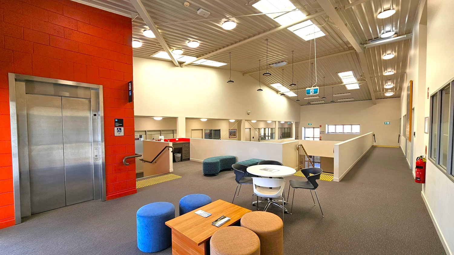 A modern indoor space with a grey carpeted floor. On the left, there's a silver lift set in a red brick wall. In the centre, a seating area features a round white table surrounded by black and white chairs, and several colourful pouffes. A wooden table holds some pamphlets. The right side has a corridor with a fire extinguisher on the wall. The ceiling is high with exposed beams and multiple lights, and a staircase is visible in the background. A modern indoor space with a grey carpeted floor. On the left, there's a silver lift set in a red brick wall. In the centre, a seating area features a round white table surrounded by black and white chairs, and several colourful pouffes. A wooden table holds some pamphlets. The right side has a corridor with a fire extinguisher on the wall. The ceiling is high with exposed beams and multiple lights, and a staircase is visible in the background.