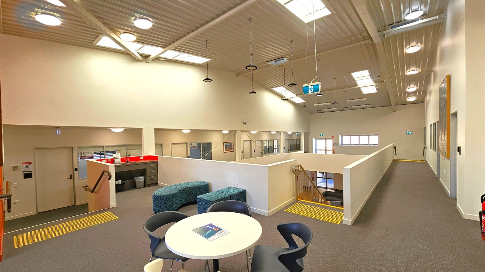 A spacious interior with a high, corrugated metal ceiling featuring multiple skylights and hanging lights. In the foreground, a round table with chairs and a notebook is visible. To the left, a small kitchenette area with a sink and cabinets is adjacent to a staircase with a handrail. The right side has a long corridor with doors and a fire extinguisher. The floor is carpeted, and there are yellow tactile indicators near the stairs. Walls are light-coloured, enhancing the bright atmosphere. A spacious interior with a high, corrugated metal ceiling featuring multiple skylights and hanging lights. In the foreground, a round table with chairs and a notebook is visible. To the left, a small kitchenette area with a sink and cabinets is adjacent to a staircase with a handrail. The right side has a long corridor with doors and a fire extinguisher. The floor is carpeted, and there are yellow tactile indicators near the stairs. Walls are light-coloured, enhancing the bright atmosphere.