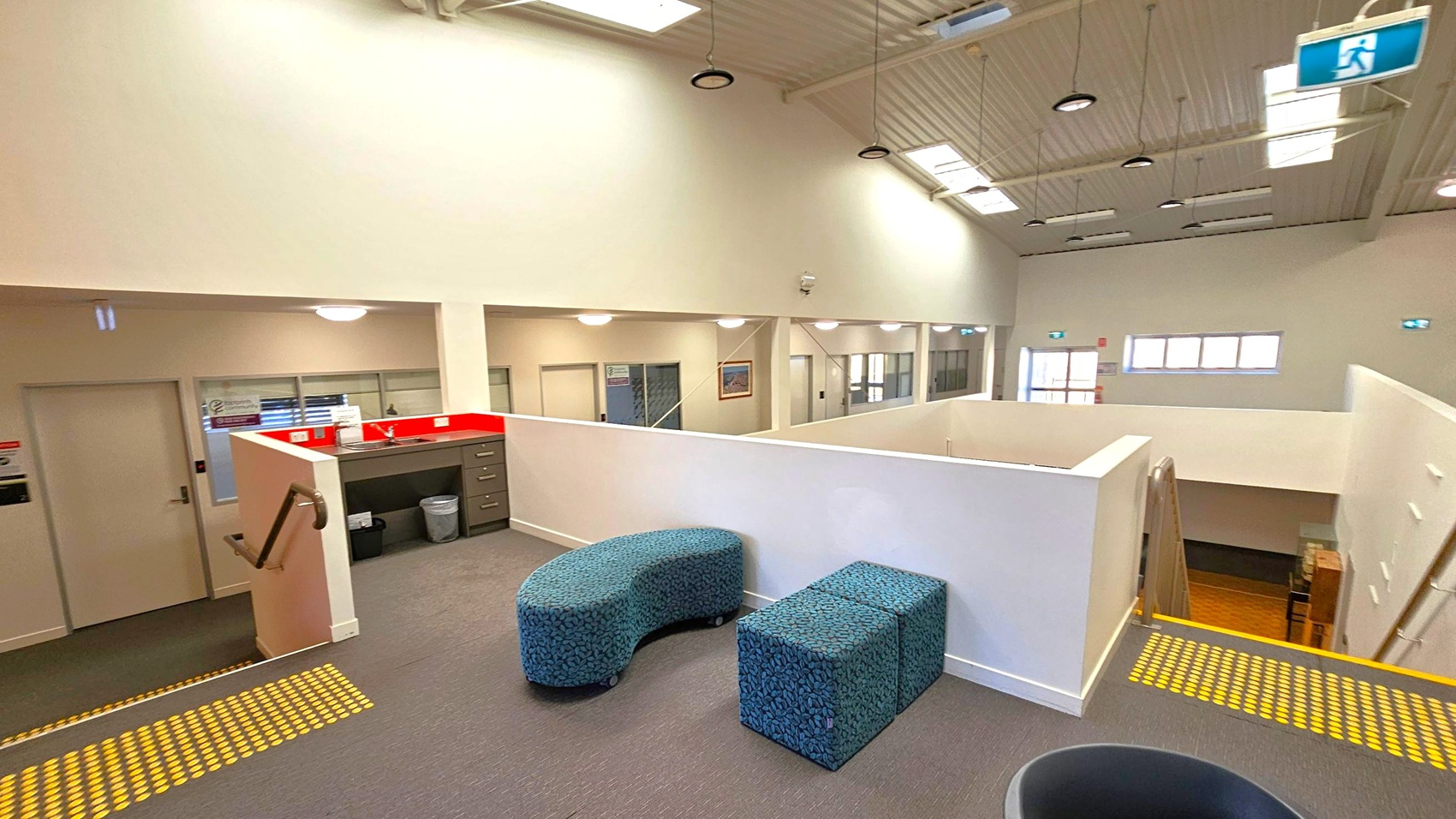 A spacious interior with a high ceiling featuring skylights and hanging lights. In the foreground, there are two blue, patterned seating units on a grey carpet. To the left, a small kitchenette with a red splashback and cabinets. A staircase with a handrail descends to the lower level. The background shows a corridor with multiple doors and windows, and a few framed pictures on the walls. An emergency exit sign is visible near the ceiling. A spacious interior with a high ceiling featuring skylights and hanging lights. In the foreground, there are two blue, patterned seating units on a grey carpet. To the left, a small kitchenette with a red splashback and cabinets. A staircase with a handrail descends to the lower level. The background shows a corridor with multiple doors and windows, and a few framed pictures on the walls. An emergency exit sign is visible near the ceiling.