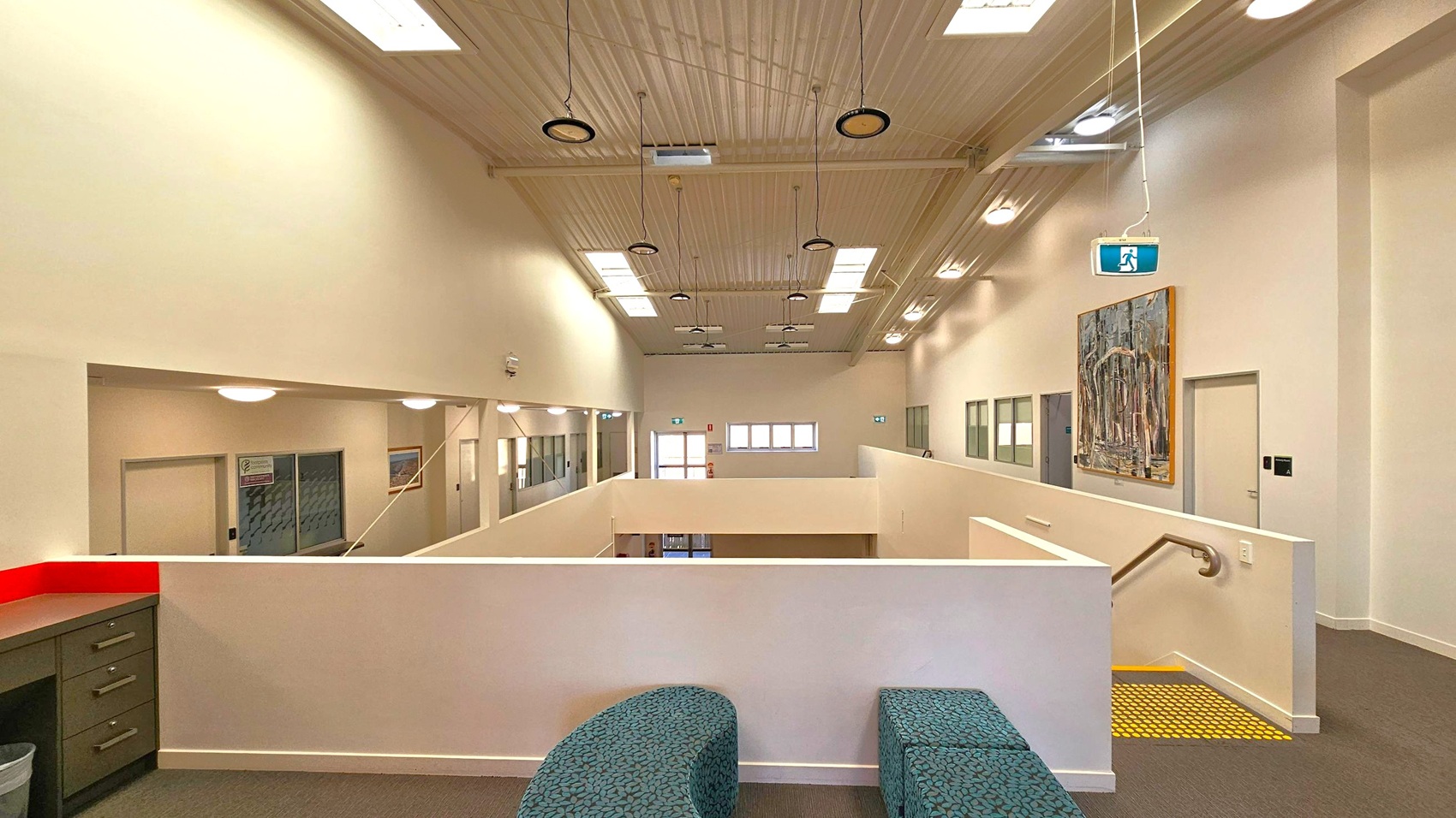 A spacious interior with a high, wooden-panelled ceiling featuring multiple skylights and hanging lights. The foreground has a curved, patterned bench and a green cabinet with drawers. The background shows a corridor with doors and windows, a large painting on the right wall, and an exit sign hanging from the ceiling. The floor is carpeted, and a staircase with a yellow safety strip is visible on the right. A spacious interior with a high, wooden-panelled ceiling featuring multiple skylights and hanging lights. The foreground has a curved, patterned bench and a green cabinet with drawers. The background shows a corridor with doors and windows, a large painting on the right wall, and an exit sign hanging from the ceiling. The floor is carpeted, and a staircase with a yellow safety strip is visible on the right.