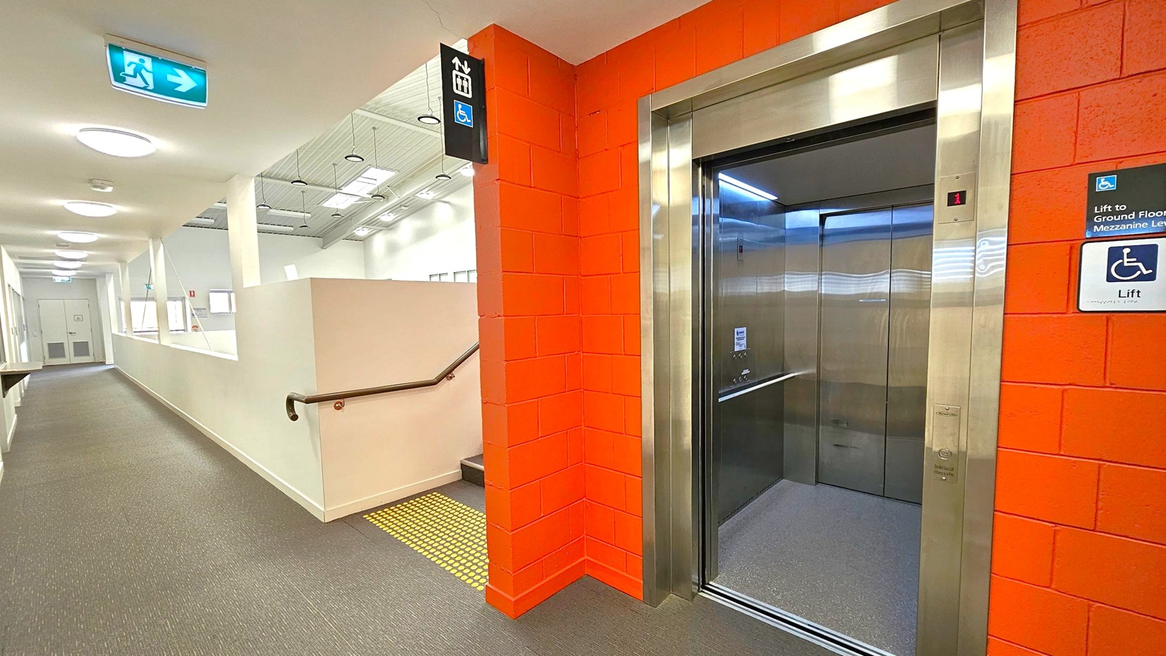 A corridor with a grey carpeted floor and white walls. On the right, there's an open lift with a stainless steel frame set into a red brick wall. Above the lift, a sign indicates accessibility. To the left, a series of round ceiling lights illuminate the hallway, which leads to a door at the end. An emergency exit sign with a running figure and arrow is mounted on the ceiling. A handrail runs along the wall, and a small staircase is visible near the lift. A corridor with a grey carpeted floor and white walls. On the right, there's an open lift with a stainless steel frame set into a red brick wall. Above the lift, a sign indicates accessibility. To the left, a series of round ceiling lights illuminate the hallway, which leads to a door at the end. An emergency exit sign with a running figure and arrow is mounted on the ceiling. A handrail runs along the wall, and a small staircase is visible near the lift.