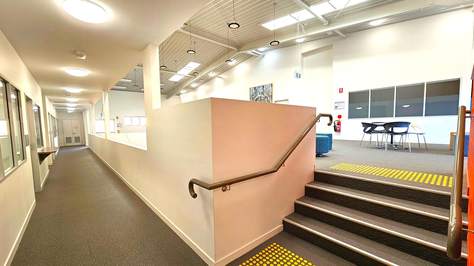 A corridor with a brown carpeted floor on the left, featuring a row of ceiling lights and windows along the wall. On the right, a short flight of stairs with a handrail leads up to a carpeted area with a table and chairs. The ceiling is high with exposed beams and hanging lights. Skylights allow natural light in. A fire extinguisher is mounted on the wall near the stairs, and a piece of artwork is displayed above a blue cushioned seat. A corridor with a brown carpeted floor on the left, featuring a row of ceiling lights and windows along the wall. On the right, a short flight of stairs with a handrail leads up to a carpeted area with a table and chairs. The ceiling is high with exposed beams and hanging lights. Skylights allow natural light in. A fire extinguisher is mounted on the wall near the stairs, and a piece of artwork is displayed above a blue cushioned seat.