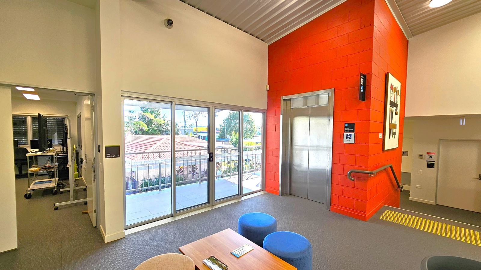 A room with a grey carpeted floor and a corrugated metal ceiling. On the right, there's a red brick wall with a lift and a handrail leading to stairs. A piece of abstract art is mounted on the wall. In the centre, a set of large glass sliding doors opens to a balcony with a view of trees and rooftops. On the left, an open doorway leads to a room with computer equipment. In the foreground, a wooden table with brochures and two blue stools are visible. A room with a grey carpeted floor and a corrugated metal ceiling. On the right, there's a red brick wall with a lift and a handrail leading to stairs. A piece of abstract art is mounted on the wall. In the centre, a set of large glass sliding doors opens to a balcony with a view of trees and rooftops. On the left, an open doorway leads to a room with computer equipment. In the foreground, a wooden table with brochures and two blue stools are visible.