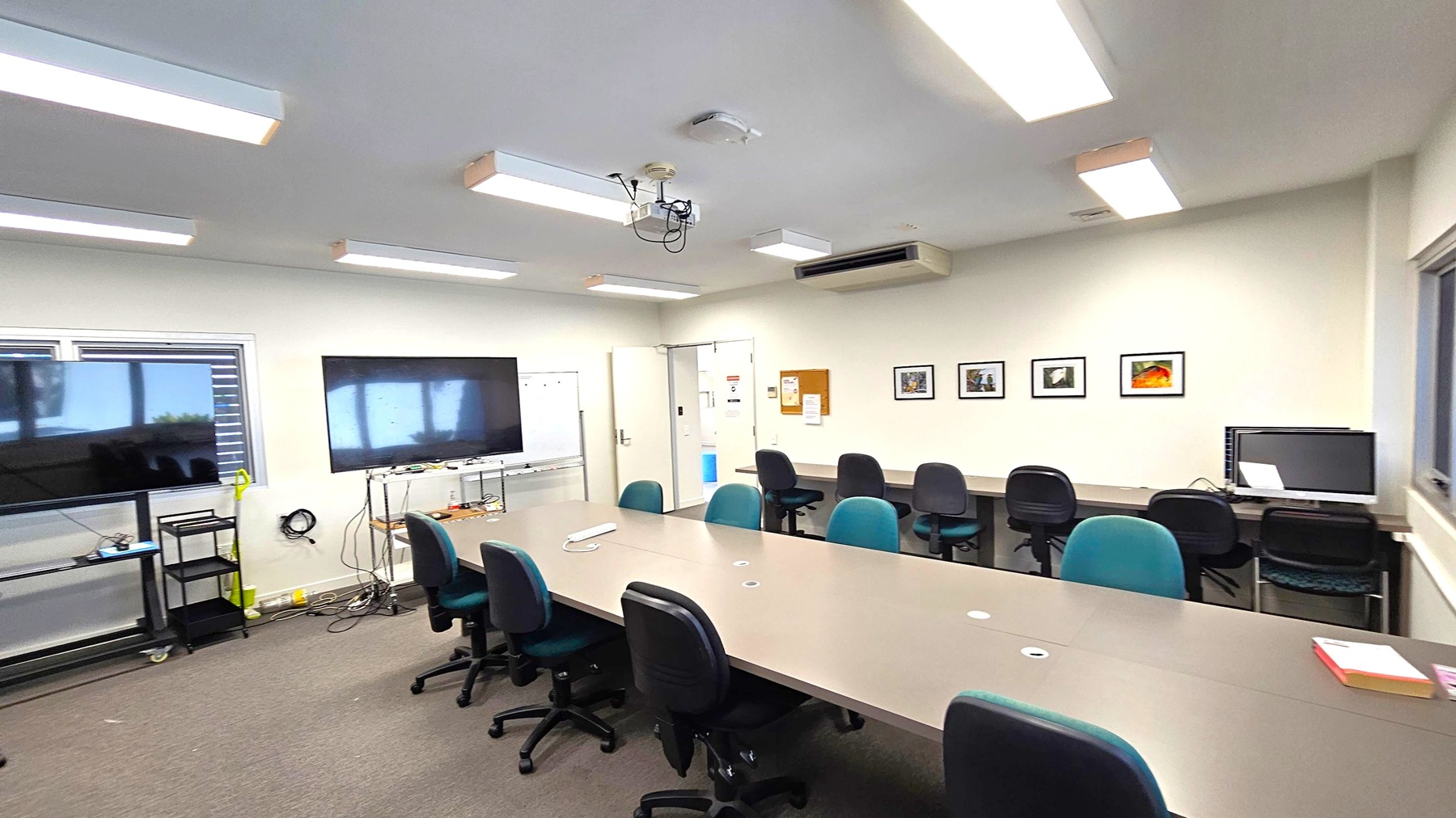 A conference room with a long table surrounded by office chairs. On the left, there are two large screens, one on a stand and another mounted on the wall, with a whiteboard nearby. The right wall features framed pictures and a computer on a desk. The ceiling has several fluorescent lights and a projector. The floor is carpeted, and there are windows with blinds on the left side. A conference room with a long table surrounded by office chairs. On the left, there are two large screens, one on a stand and another mounted on the wall, with a whiteboard nearby. The right wall features framed pictures and a computer on a desk. The ceiling has several fluorescent lights and a projector. The floor is carpeted, and there are windows with blinds on the left side.