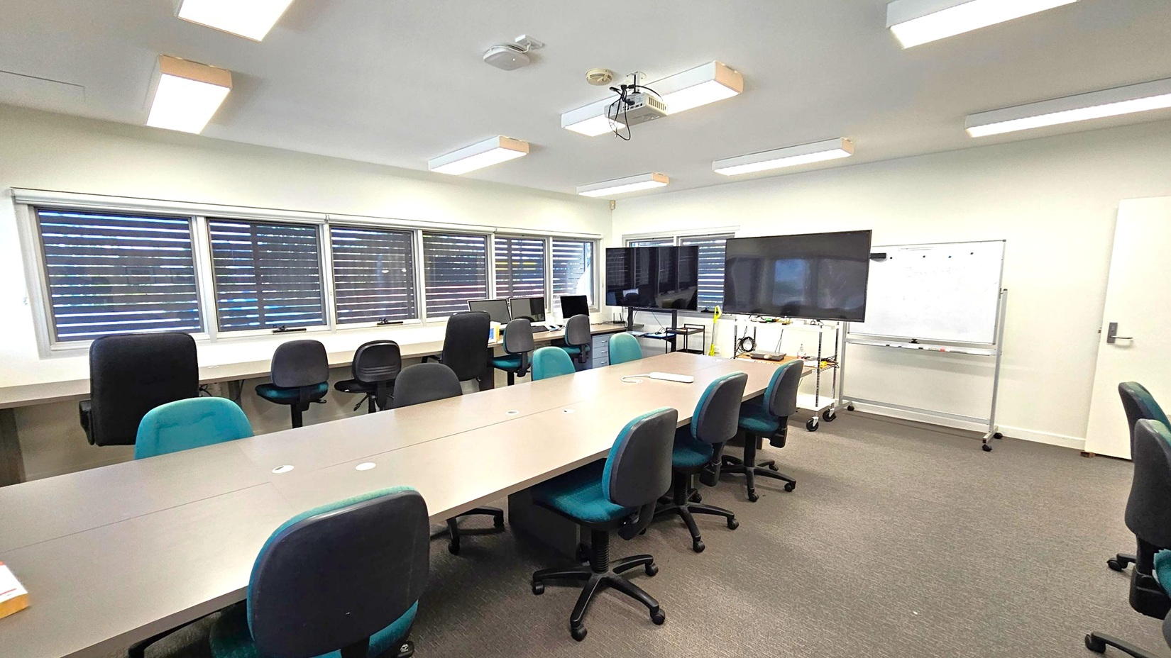 A conference room with a long table in the centre surrounded by office chairs. On the left, there are windows with blinds and a row of desks with computers and chairs. On the right, a large monitor is mounted on a stand, and a whiteboard is nearby. The ceiling has several fluorescent lights and a projector. The floor is carpeted in a brown and green pattern. A conference room with a long table in the centre surrounded by office chairs. On the left, there are windows with blinds and a row of desks with computers and chairs. On the right, a large monitor is mounted on a stand, and a whiteboard is nearby. The ceiling has several fluorescent lights and a projector. The floor is carpeted in a brown and green pattern.