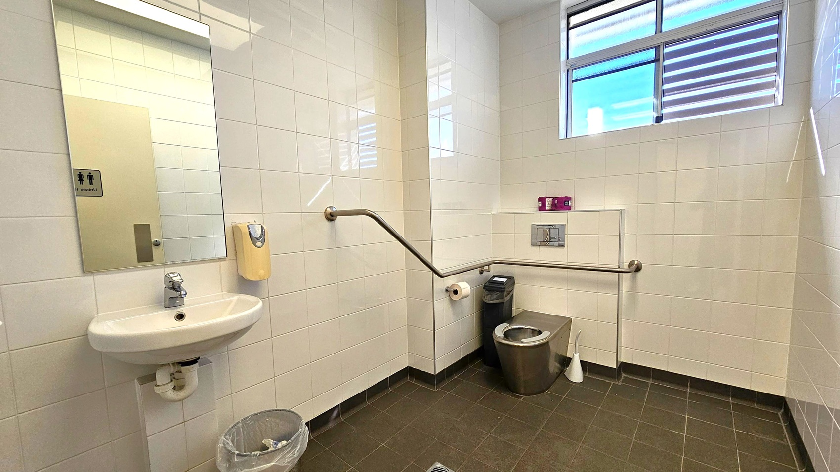 A public restroom with white tiled walls and a dark tiled floor. On the left, there's a sink with a mirror above it and a soap dispenser mounted on the wall. Below the sink is a small waste bin. On the right, a stainless steel toilet with a toilet roll holder and a sanitary bin nearby. A grab bar runs along the wall for accessibility. Above, a window with horizontal blinds allows natural light into the room. A public restroom with white tiled walls and a dark tiled floor. On the left, there's a sink with a mirror above it and a soap dispenser mounted on the wall. Below the sink is a small waste bin. On the right, a stainless steel toilet with a toilet roll holder and a sanitary bin nearby. A grab bar runs along the wall for accessibility. Above, a window with horizontal blinds allows natural light into the room.