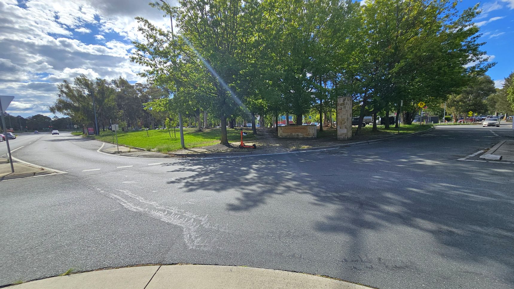 A street intersection with a road curving to the left and another road extending to the right. In the centre, there are several large trees providing shade, with a sign partially visible among them. A scooter is parked near the trees. The pavement is smooth, and the area is well-lit with sunlight filtering through the leaves. On the left, a grassy area is bordered by a footpath, and a few cars are visible in the distance. A street intersection with a road curving to the left and another road extending to the right. In the centre, there are several large trees providing shade, with a sign partially visible among them. A scooter is parked near the trees. The pavement is smooth, and the area is well-lit with sunlight filtering through the leaves. On the left, a grassy area is bordered by a footpath, and a few cars are visible in the distance.