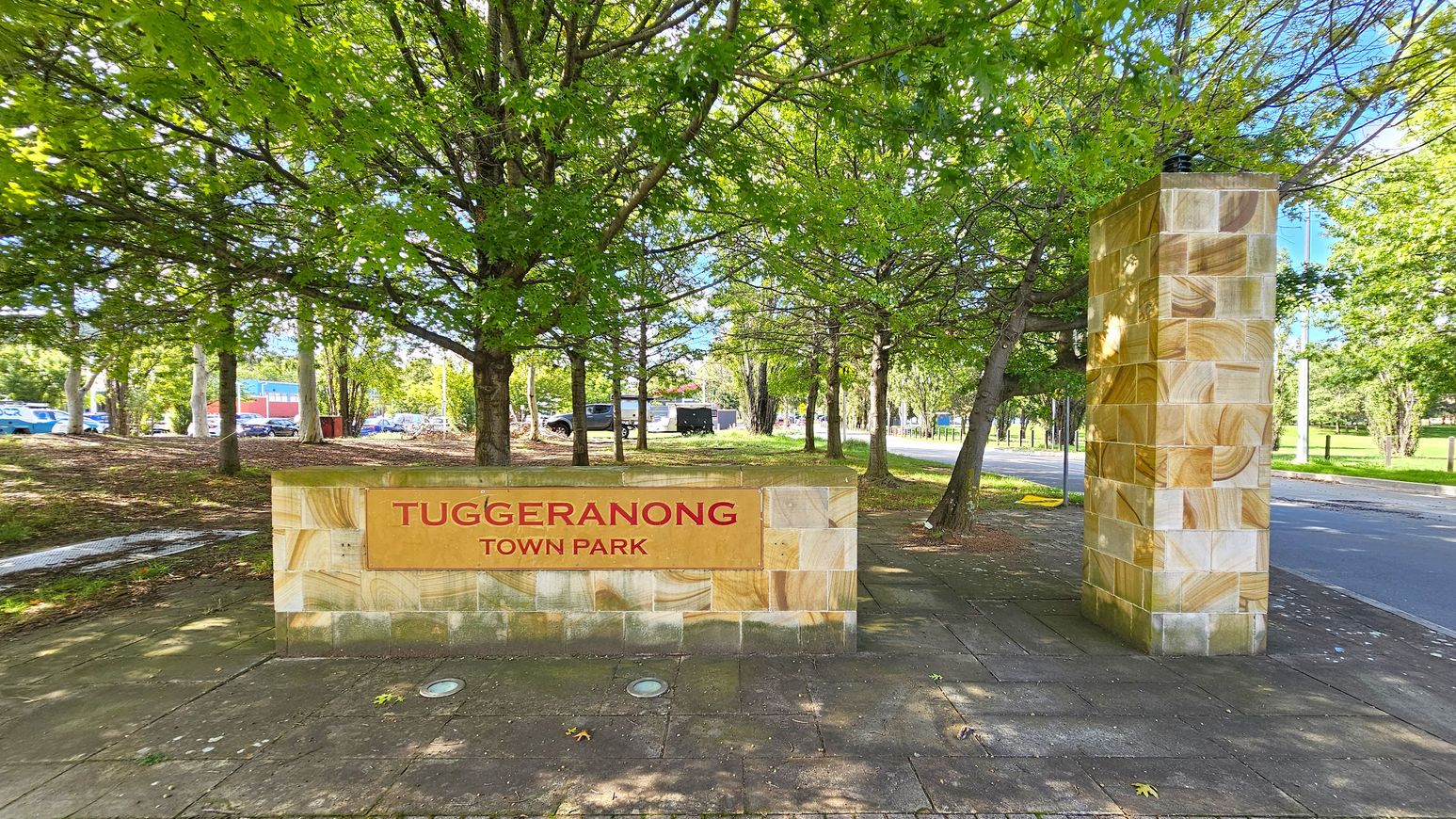 A stone sign with "Tuggeranong Town Park" in red letters is on the left, set against a backdrop of lush green trees. To the right, a tall stone pillar stands beside a paved path. The ground is covered with square tiles, and a road runs parallel to the path. In the background, more trees and a few parked cars are visible. A stone sign with "Tuggeranong Town Park" in red letters is on the left, set against a backdrop of lush green trees. To the right, a tall stone pillar stands beside a paved path. The ground is covered with square tiles, and a road runs parallel to the path. In the background, more trees and a few parked cars are visible.