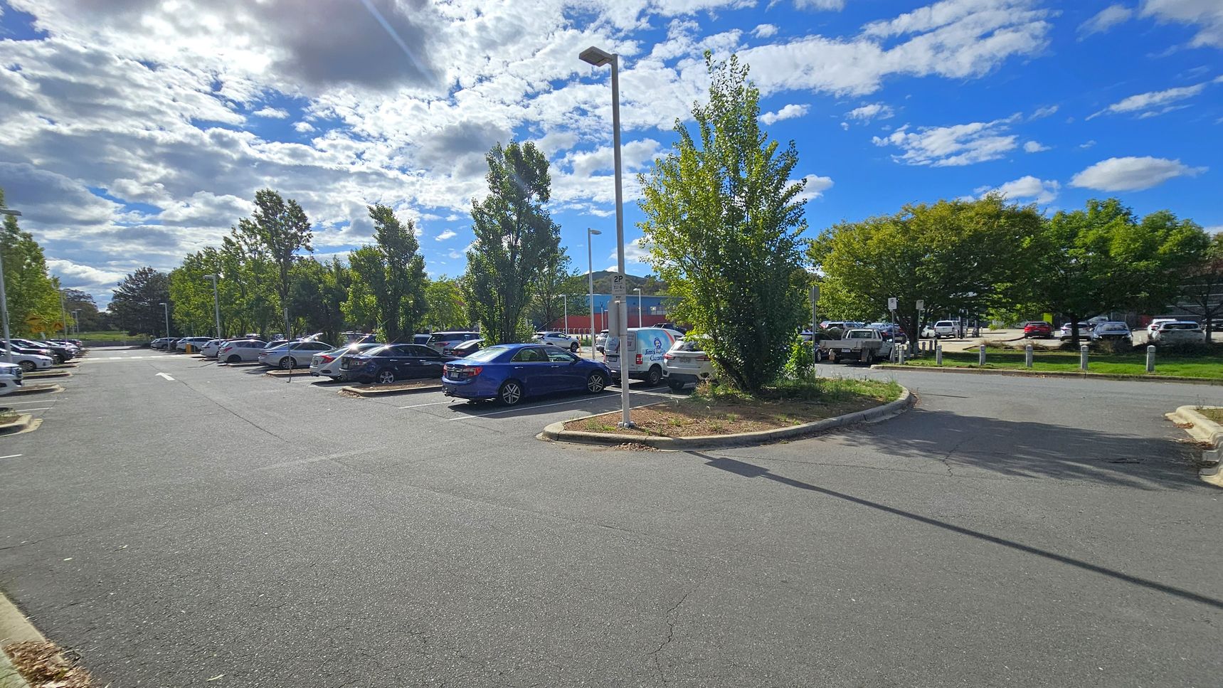 A car park with several vehicles is visible, surrounded by trees. On the left, there are rows of parked cars and tall streetlights. In the centre, a blue car is parked near a small island with a tree and a signpost. On the right, more cars are parked, and a building with a blue and red facade is partially visible in the background. The ground is paved with asphalt, and the area is well-lit by daylight. A car park with several vehicles is visible, surrounded by trees. On the left, there are rows of parked cars and tall streetlights. In the centre, a blue car is parked near a small island with a tree and a signpost. On the right, more cars are parked, and a building with a blue and red facade is partially visible in the background. The ground is paved with asphalt, and the area is well-lit by daylight.