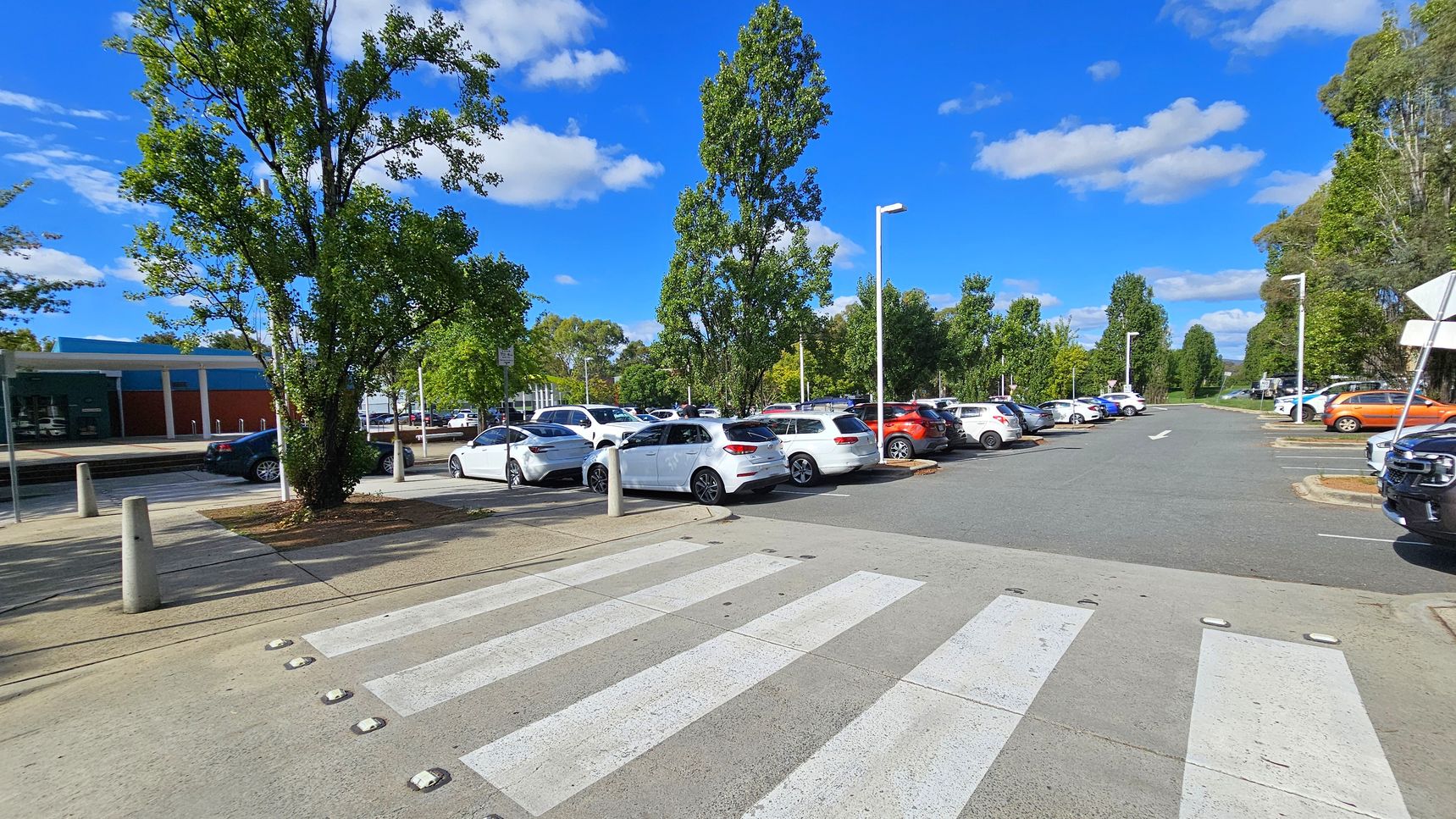 A car park with a zebra crossing in the foreground. On the left, there is a large tree and a building with a blue and red facade. Several cars are parked, mostly white, with a few in other colours like orange. Tall trees line the parking area, and street lamps are visible. The road extends into the distance, flanked by more trees and greenery. The sky is clear with a few clouds. A car park with a zebra crossing in the foreground. On the left, there is a large tree and a building with a blue and red facade. Several cars are parked, mostly white, with a few in other colours like orange. Tall trees line the parking area, and street lamps are visible. The road extends into the distance, flanked by more trees and greenery. The sky is clear with a few clouds.