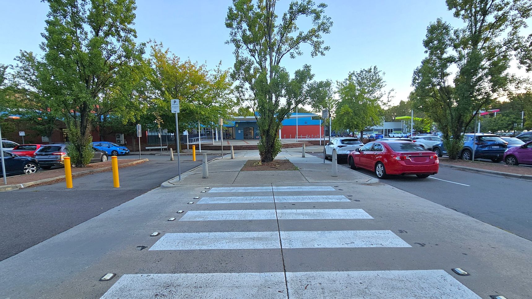 A zebra crossing leads to a tree-lined area with a central tree. On the left, there are parked cars and yellow bollards along the road. On the right, a red car is parked near more trees and additional vehicles. In the background, a building with blue and red accents is visible, surrounded by more trees and a parking area. The ground is paved, and the scene is well-lit. A zebra crossing leads to a tree-lined area with a central tree. On the left, there are parked cars and yellow bollards along the road. On the right, a red car is parked near more trees and additional vehicles. In the background, a building with blue and red accents is visible, surrounded by more trees and a parking area. The ground is paved, and the scene is well-lit.