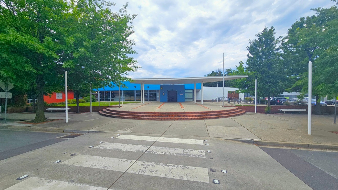 A building with a blue and white facade is in the centre, featuring a circular entrance area with steps leading up to it. The foreground has a zebra crossing on a paved surface. On the left and right, there are large green trees and white lamp posts. The area is open and spacious, with a few benches visible near the trees. A building with a blue and white facade is in the centre, featuring a circular entrance area with steps leading up to it. The foreground has a zebra crossing on a paved surface. On the left and right, there are large green trees and white lamp posts. The area is open and spacious, with a few benches visible near the trees.
