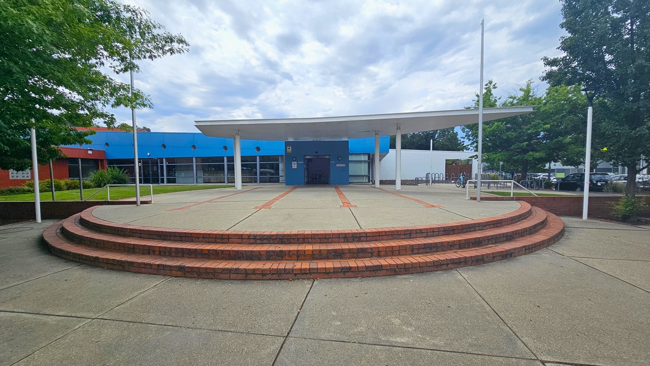 A circular, tiered brick platform is in the foreground, leading up to a covered entrance. The building has a blue facade with large windows and a flat roof supported by white pillars. On the left, there are green trees and a red section of the building. On the right, more trees and a glimpse of parked cars are visible. The ground is paved with concrete, and the sky is overcast. A circular, tiered brick platform is in the foreground, leading up to a covered entrance. The building has a blue facade with large windows and a flat roof supported by white pillars. On the left, there are green trees and a red section of the building. On the right, more trees and a glimpse of parked cars are visible. The ground is paved with concrete, and the sky is overcast.