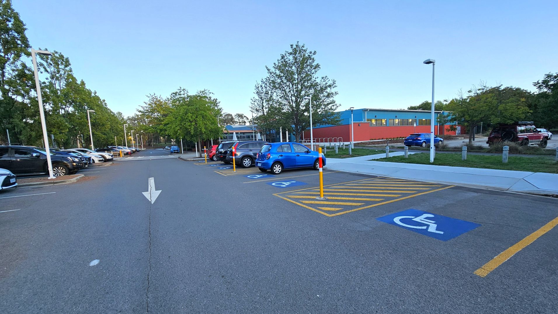 A car park with a row of parked cars on the left, including black and silver vehicles. On the right, there are designated disabled parking spaces marked with blue signs and yellow lines. A blue car is parked in one of these spaces. In the background, a building with blue and orange walls is visible, surrounded by trees. The ground is paved, and there are streetlights along the parking area. An arrow is painted on the road, indicating direction. A car park with a row of parked cars on the left, including black and silver vehicles. On the right, there are designated disabled parking spaces marked with blue signs and yellow lines. A blue car is parked in one of these spaces. In the background, a building with blue and orange walls is visible, surrounded by trees. The ground is paved, and there are streetlights along the parking area. An arrow is painted on the road, indicating direction.