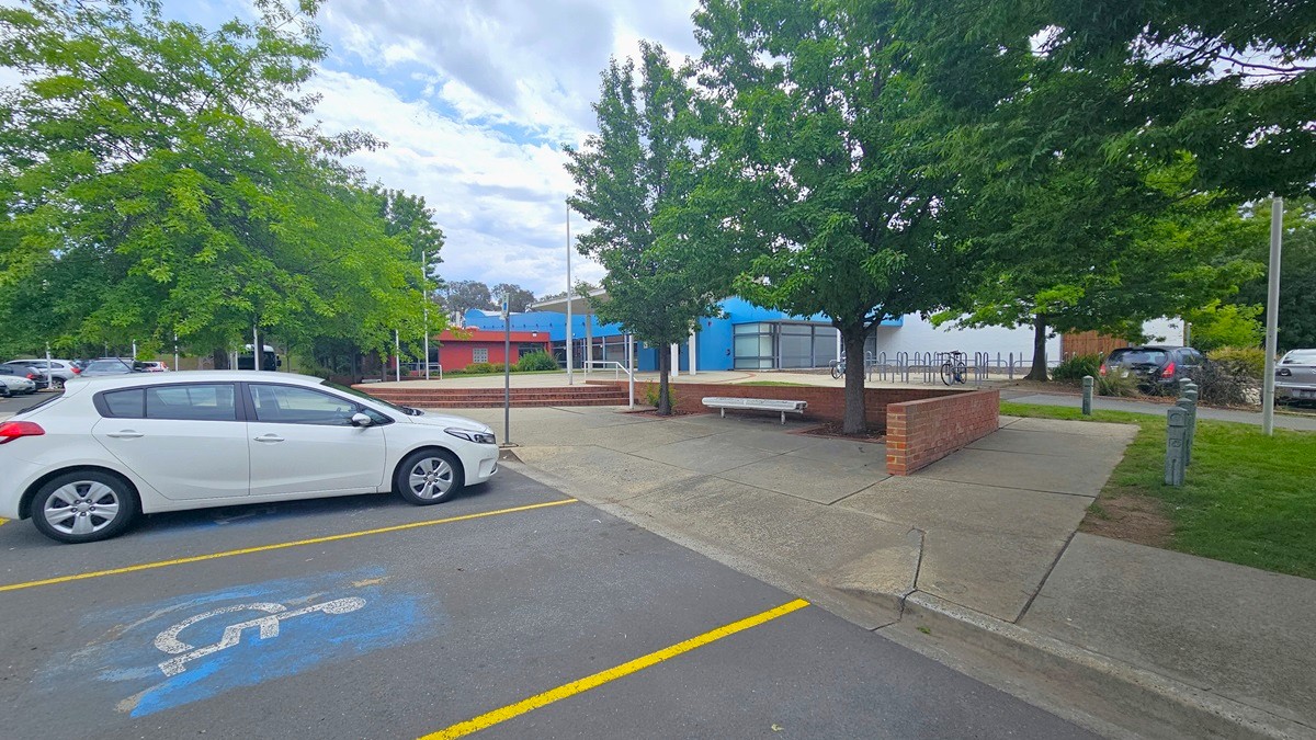 A white car is parked in a designated disabled parking space on the left, marked by blue paint and a wheelchair symbol. The ground is paved with yellow lines marking the parking area. In the middle, there are trees providing shade, and a brick bench is visible. In the background, a building with blue and red accents is partially obscured by trees. Additional cars are parked further back, and a bicycle rack is visible near the building. A white car is parked in a designated disabled parking space on the left, marked by blue paint and a wheelchair symbol. The ground is paved with yellow lines marking the parking area. In the middle, there are trees providing shade, and a brick bench is visible. In the background, a building with blue and red accents is partially obscured by trees. Additional cars are parked further back, and a bicycle rack is visible near the building.