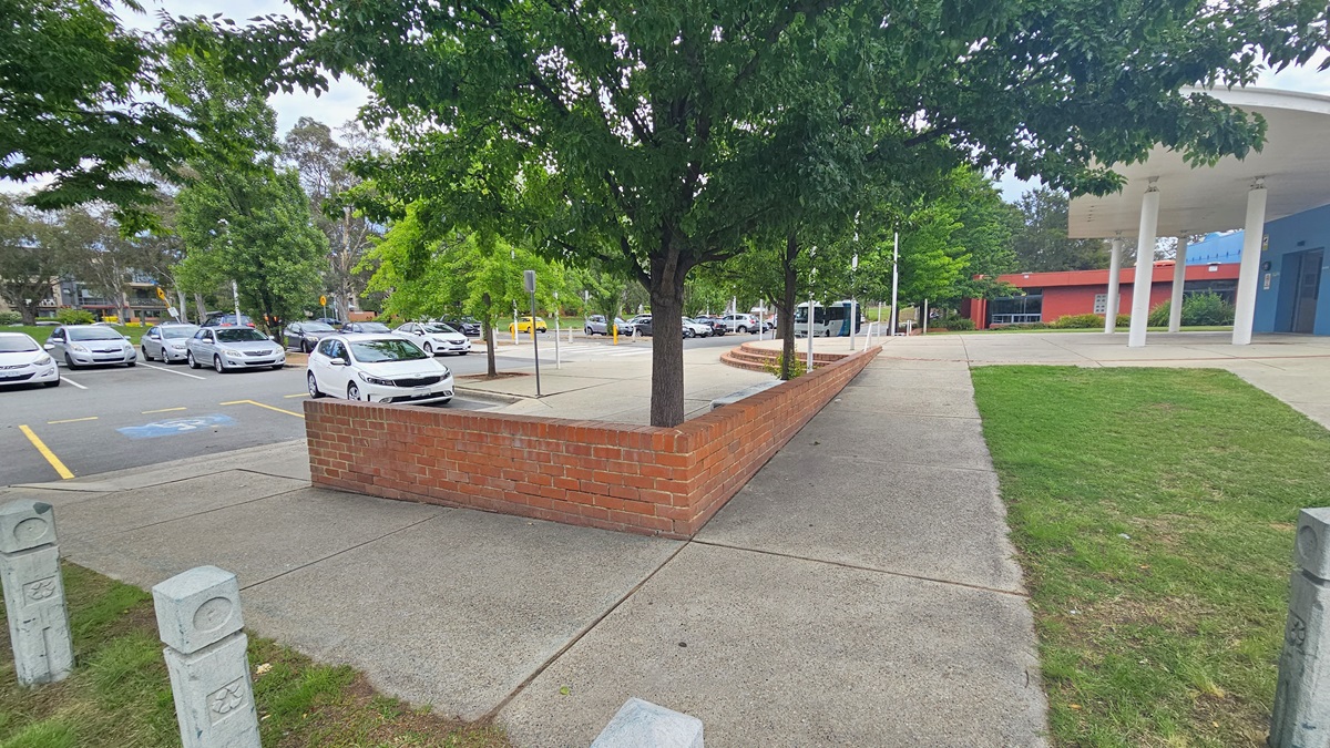 A paved area with a red brick wall runs through the centre, with a tree growing from it. On the left, there is a car park with several parked cars and yellow lines marking spaces. On the right, a grassy area leads to a building with a blue and red facade supported by white columns. The background features more trees and additional parked cars. The ground is a mix of concrete and grass. A paved area with a red brick wall runs through the centre, with a tree growing from it. On the left, there is a car park with several parked cars and yellow lines marking spaces. On the right, a grassy area leads to a building with a blue and red facade supported by white columns. The background features more trees and additional parked cars. The ground is a mix of concrete and grass.