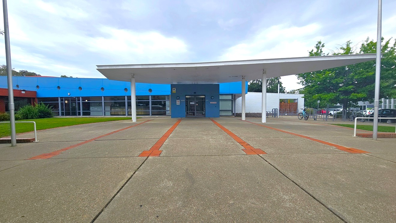 A building with a blue facade and a large white overhang supported by pillars. The entrance is centred, with glass doors and windows. The ground is paved with concrete, featuring red brick lines. On the left, there's a grassy area with shrubs, and on the right, trees and a bicycle rack. The background includes parked cars and more greenery. A building with a blue facade and a large white overhang supported by pillars. The entrance is centred, with glass doors and windows. The ground is paved with concrete, featuring red brick lines. On the left, there's a grassy area with shrubs, and on the right, trees and a bicycle rack. The background includes parked cars and more greenery.