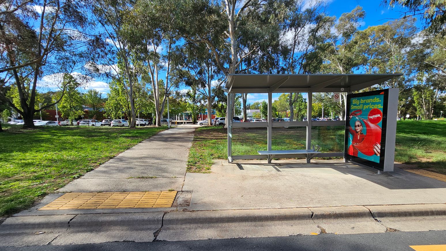 A bus stop with a metal shelter and a bench is on the right. An advertisement is displayed on the side of the shelter. A concrete path leads from the foreground to the background, flanked by grass and trees. In the background, there are parked cars and more trees. The sky is visible through the tree branches. A bus stop with a metal shelter and a bench is on the right. An advertisement is displayed on the side of the shelter. A concrete path leads from the foreground to the background, flanked by grass and trees. In the background, there are parked cars and more trees. The sky is visible through the tree branches.