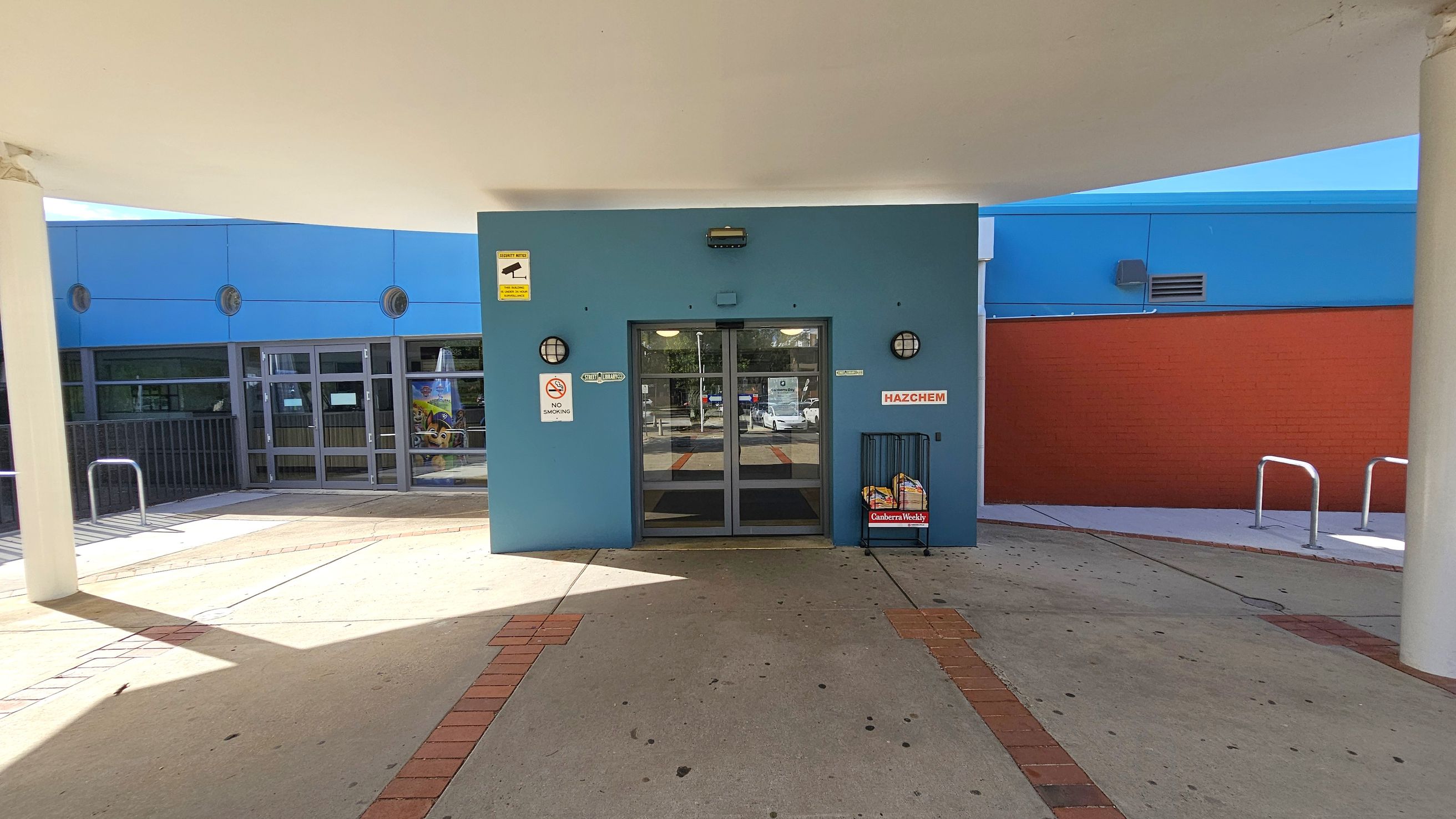 A building entrance with a blue facade and glass double doors in the centre. Above the doors, there's a security camera sign. To the left, a glass window and a metal railing are visible. On the right, a red brick wall and metal bike racks are present. A newspaper stand with "Canberra Weekly" is near the entrance. The floor is concrete with brick accents, and the ceiling is white with a curved design. A building entrance with a blue facade and glass double doors in the centre. Above the doors, there's a security camera sign. To the left, a glass window and a metal railing are visible. On the right, a red brick wall and metal bike racks are present. A newspaper stand with "Canberra Weekly" is near the entrance. The floor is concrete with brick accents, and the ceiling is white with a curved design.