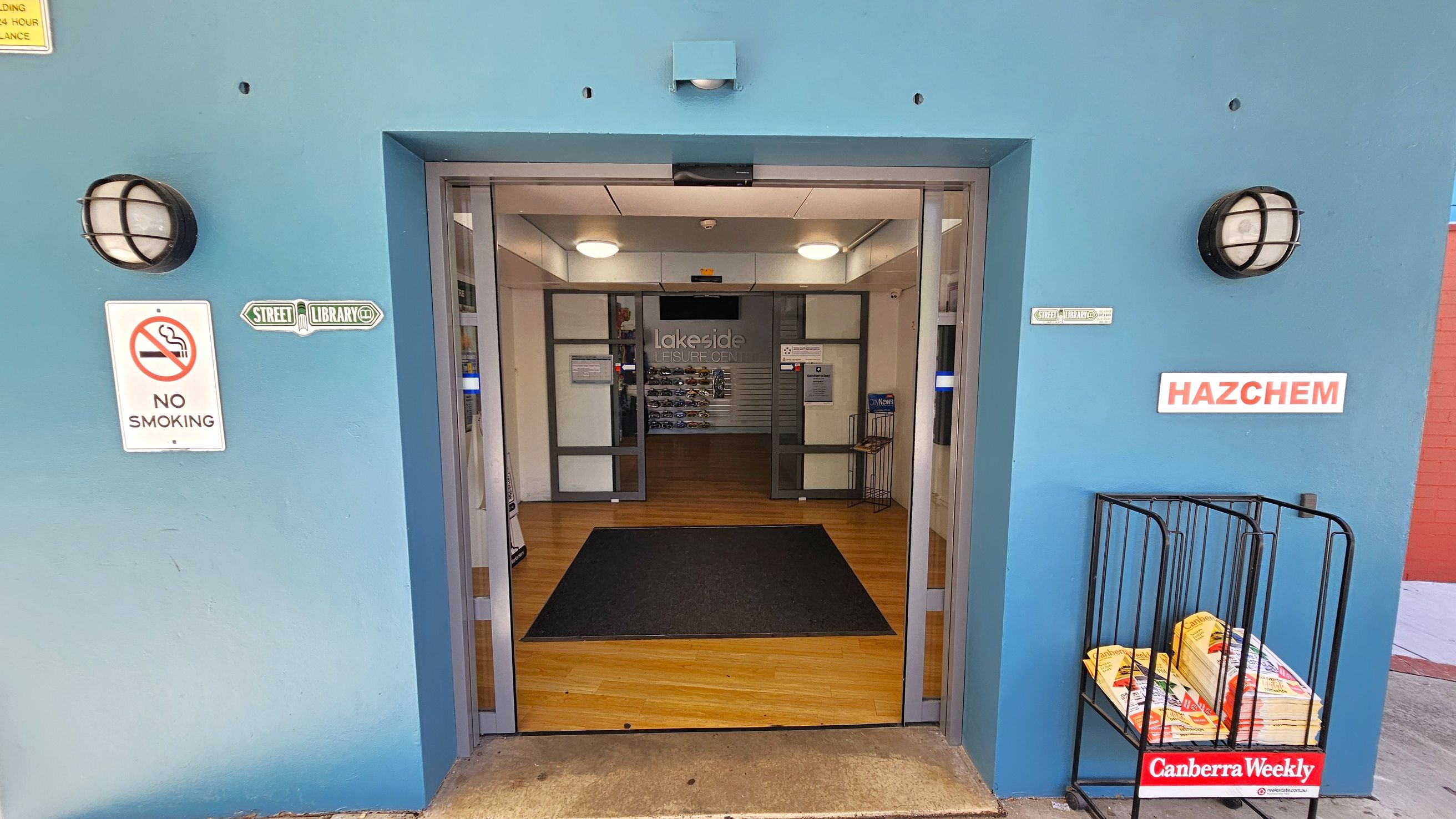 A blue entrance with double glass doors leading into a building with a wooden floor. On the left, a "No Smoking" sign and a "Street Library" sign are mounted on the wall. On the right, a "HAZCHEM" sign is visible above a rack holding copies of "Canberra Weekly". Two round wall lights are positioned on either side of the entrance. Inside, the word "Lakeside" is visible on the back wall. A blue entrance with double glass doors leading into a building with a wooden floor. On the left, a "No Smoking" sign and a "Street Library" sign are mounted on the wall. On the right, a "HAZCHEM" sign is visible above a rack holding copies of "Canberra Weekly". Two round wall lights are positioned on either side of the entrance. Inside, the word "Lakeside" is visible on the back wall.