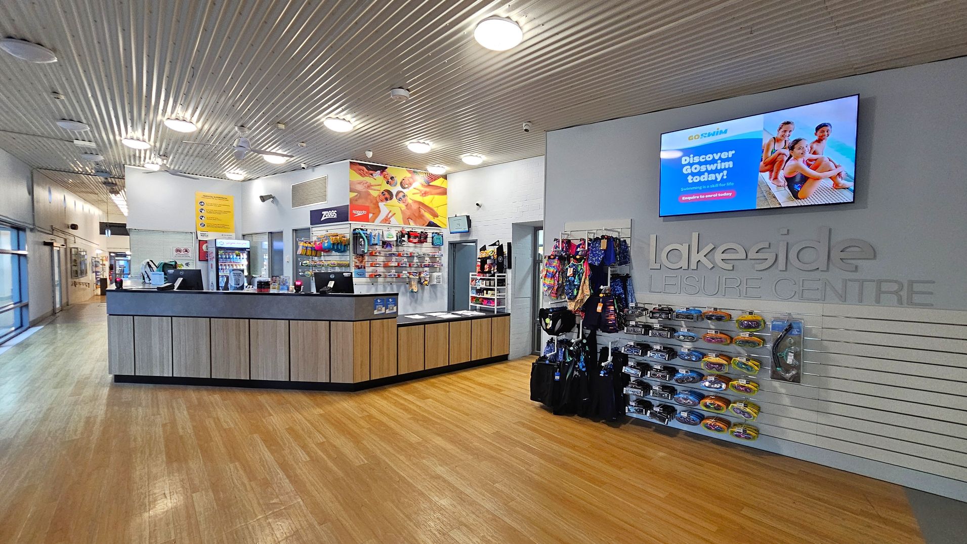 Reception area of a leisure centre with a wooden floor and a corrugated metal ceiling. On the left, a reception desk with computers and a display of swimming accessories. Behind the desk, a colourful poster and a vending machine. On the right, a wall-mounted display screen above the words "Lakeside Leisure Centre" and racks of swim gear. The background features a corridor with glass windows. Reception area of a leisure centre with a wooden floor and a corrugated metal ceiling. On the left, a reception desk with computers and a display of swimming accessories. Behind the desk, a colourful poster and a vending machine. On the right, a wall-mounted display screen above the words "Lakeside Leisure Centre" and racks of swim gear. The background features a corridor with glass windows.