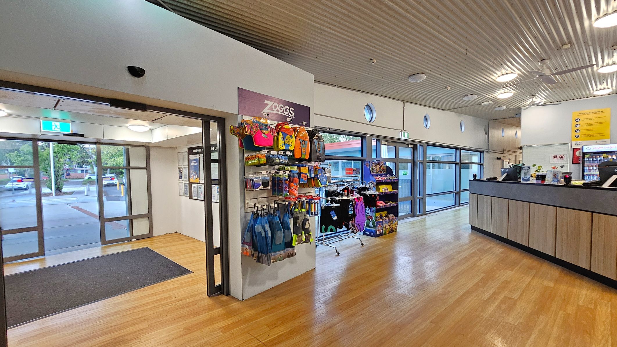 A spacious interior with a wooden floor and a corrugated metal ceiling. On the left, there is a glass entrance with a black mat and a view of parked cars outside. In the centre, a display rack features colourful swim gear and accessories. To the right, a wooden counter with a black top has a cash register and promotional materials. Large windows line the wall, allowing natural light to fill the space. A spacious interior with a wooden floor and a corrugated metal ceiling. On the left, there is a glass entrance with a black mat and a view of parked cars outside. In the centre, a display rack features colourful swim gear and accessories. To the right, a wooden counter with a black top has a cash register and promotional materials. Large windows line the wall, allowing natural light to fill the space.