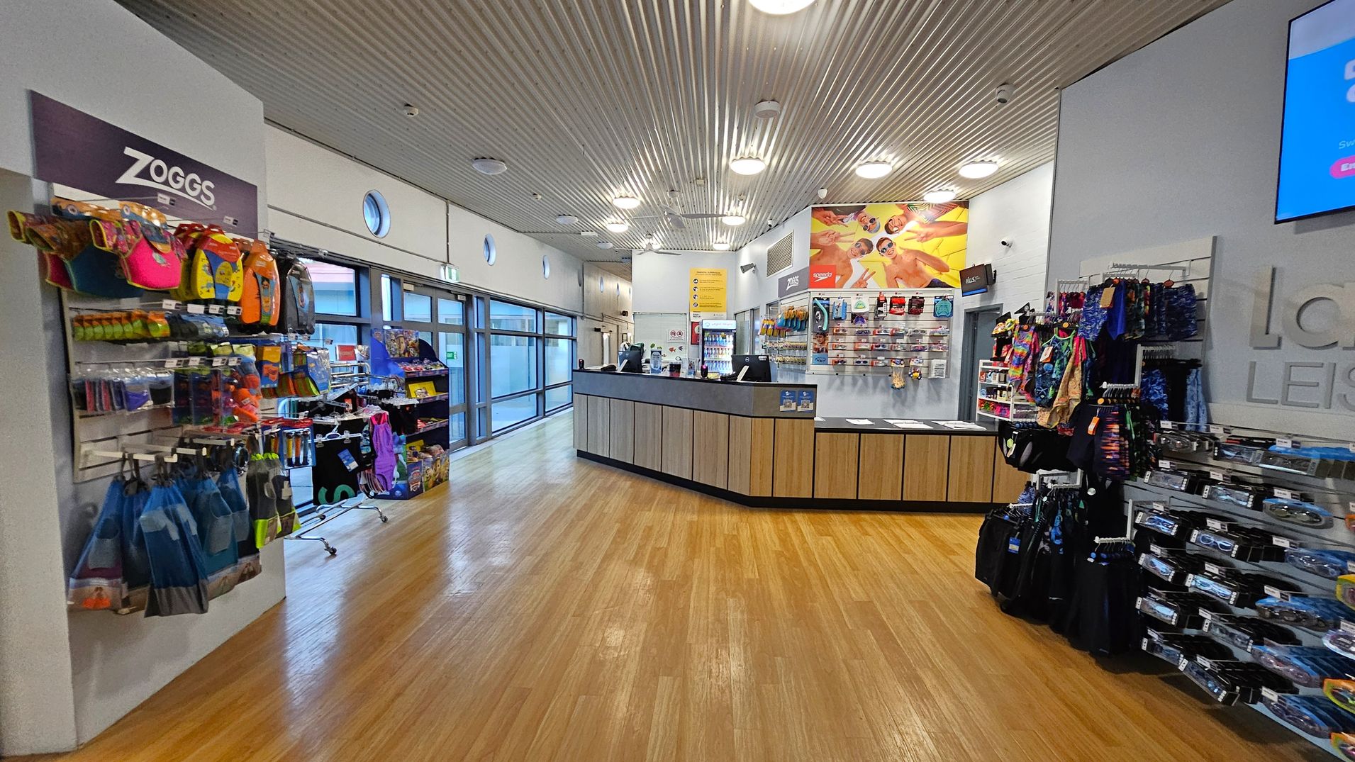 A leisure centre shop with a wooden floor and a corrugated metal ceiling. On the left, shelves display swimming gear and accessories with a "Zoggs" sign above. The centre features a reception counter with promotional materials and a display of goggles and swimwear behind it. On the right, more shelves hold colourful swimwear and accessories. Large windows line the left wall, allowing natural light to enter. A leisure centre shop with a wooden floor and a corrugated metal ceiling. On the left, shelves display swimming gear and accessories with a "Zoggs" sign above. The centre features a reception counter with promotional materials and a display of goggles and swimwear behind it. On the right, more shelves hold colourful swimwear and accessories. Large windows line the left wall, allowing natural light to enter.