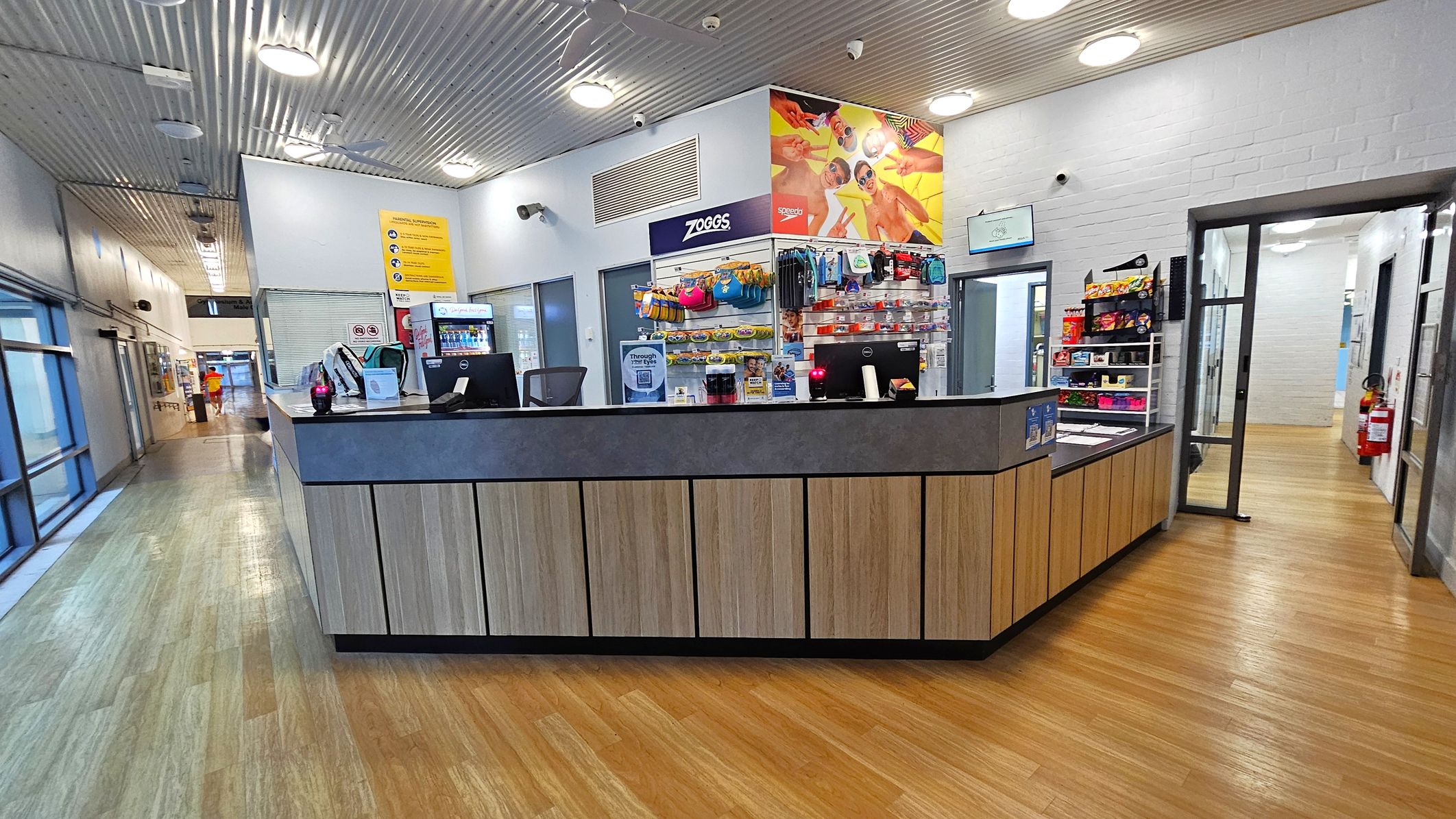 A reception area with a wooden counter in the centre, featuring two computer monitors and various items like a backpack and a drink bottle. Behind the counter, shelves display swimming gear and snacks. The floor is wooden, and the ceiling has a metallic finish with circular lights. To the left, a corridor with glass doors and windows leads to another area. On the right, a doorway opens to a hallway with a fire extinguisher mounted on the wall. A reception area with a wooden counter in the centre, featuring two computer monitors and various items like a backpack and a drink bottle. Behind the counter, shelves display swimming gear and snacks. The floor is wooden, and the ceiling has a metallic finish with circular lights. To the left, a corridor with glass doors and windows leads to another area. On the right, a doorway opens to a hallway with a fire extinguisher mounted on the wall.